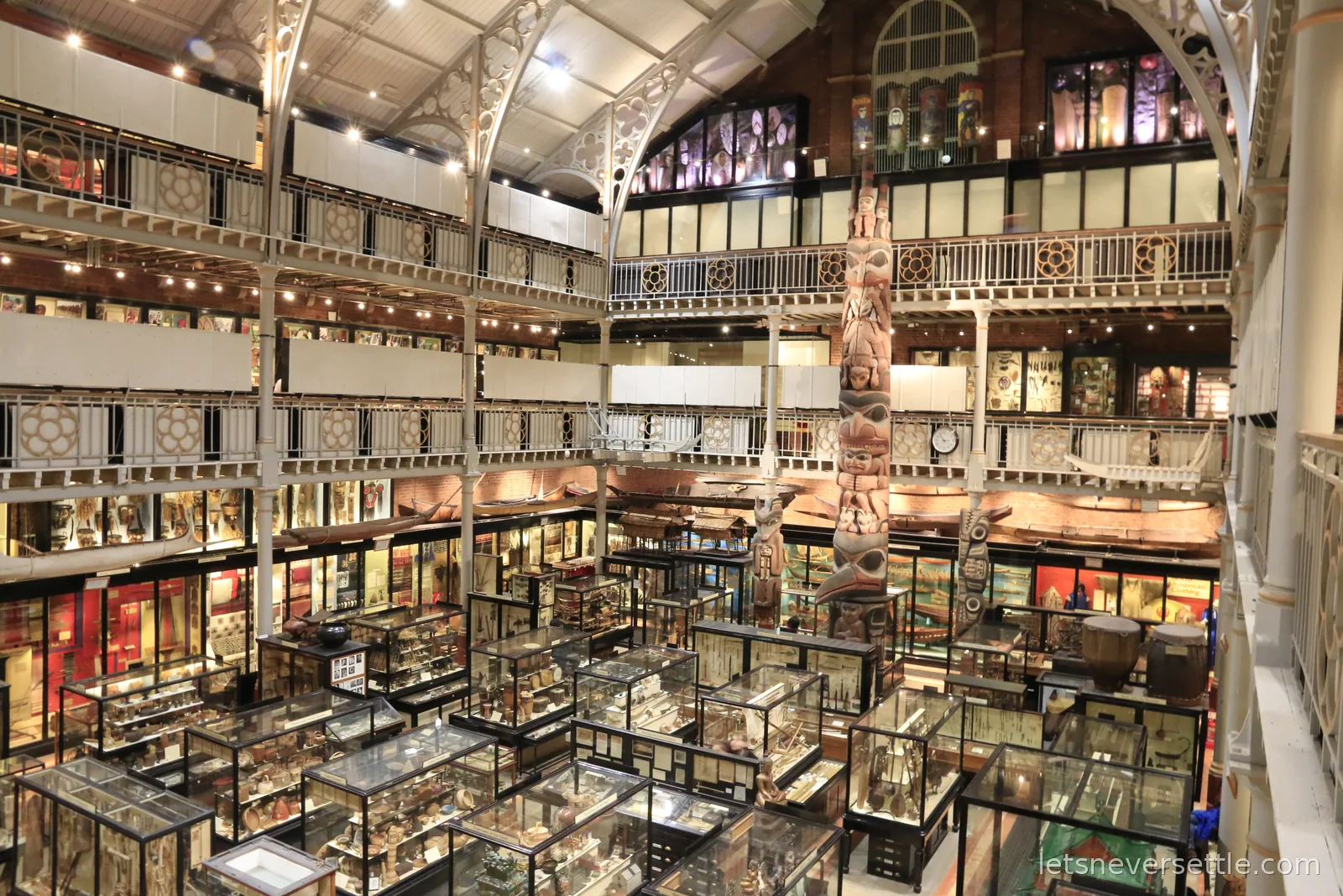 The dim interior of the Pitt Rivers Museum in Oxford, dense glass cases on two levels with a tall carved totem pole rising through the gallery