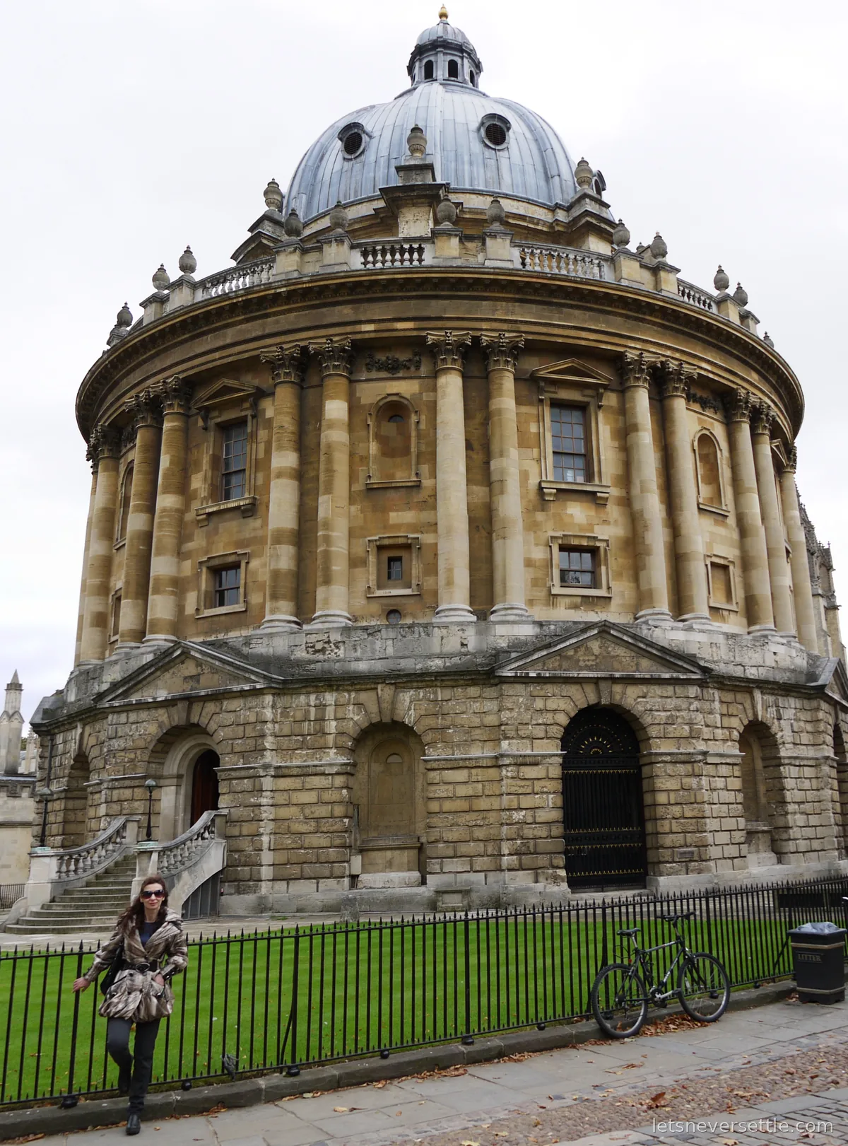Inga standing at the railings in front of the round domed Radcliffe Camera in Oxford, a bicycle leaning on the fence nearby