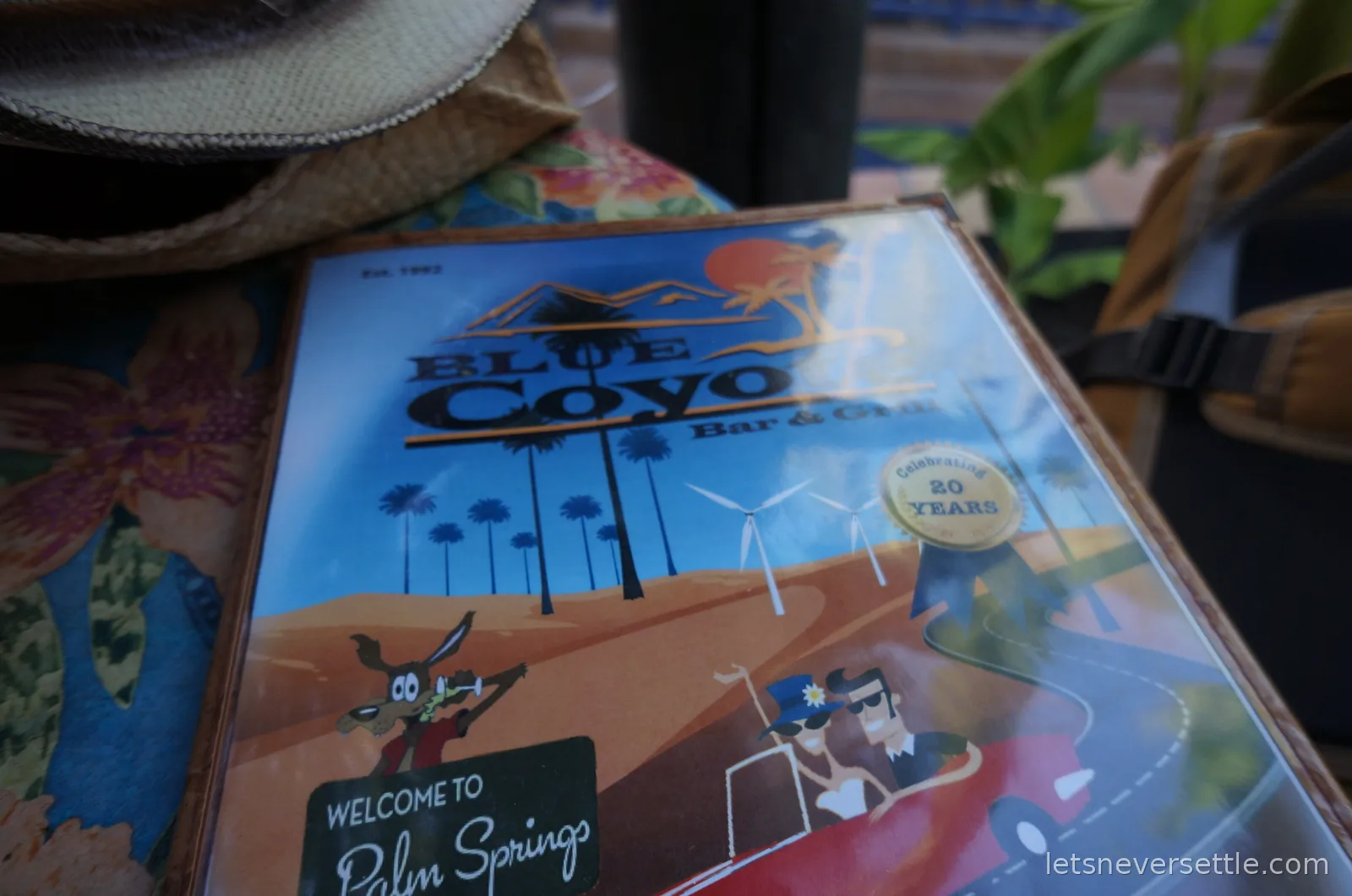 Blue Coyote Bar and Grill menu cover with a cartoon coyote and a Welcome to Palm Springs sign, a straw hat resting at the table edge