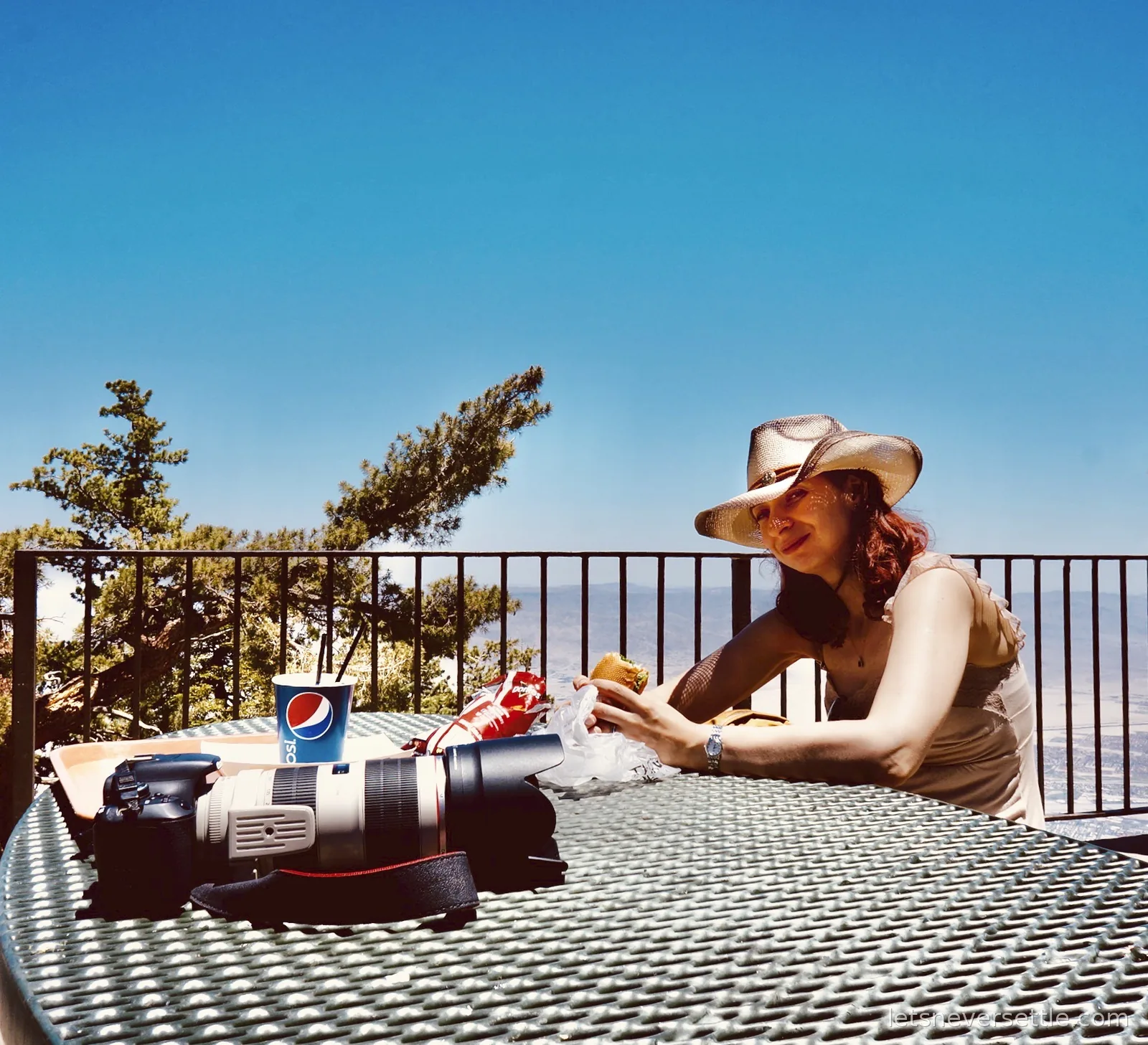 Inga in a straw hat eating a sandwich at a mountain station picnic table, camera and long lens on the table and valley haze beyond the railing