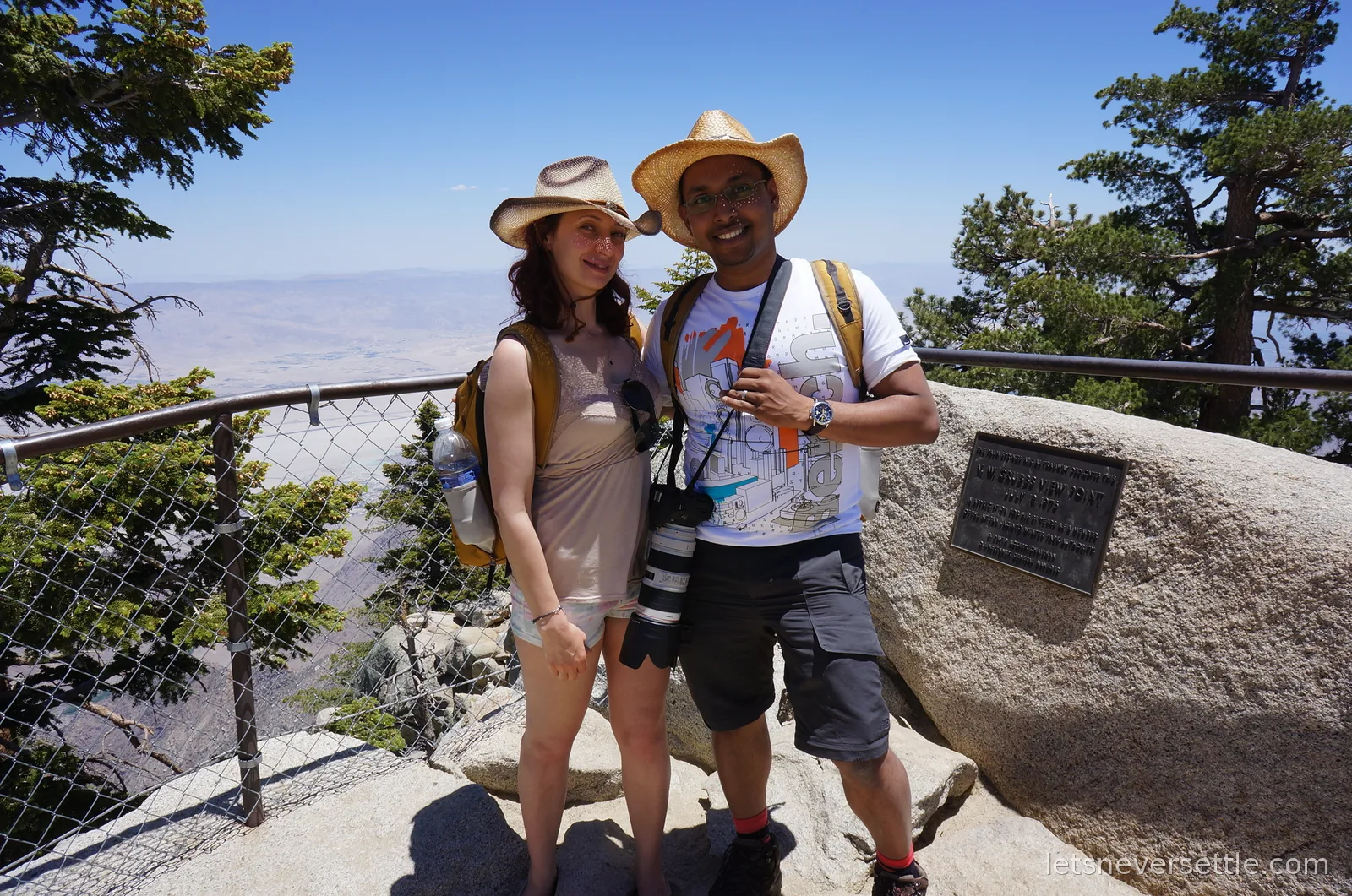 Inga and Dileep in straw hats at a fenced viewpoint on Mount San Jacinto, a plaque on a boulder beside them and the desert floor far below