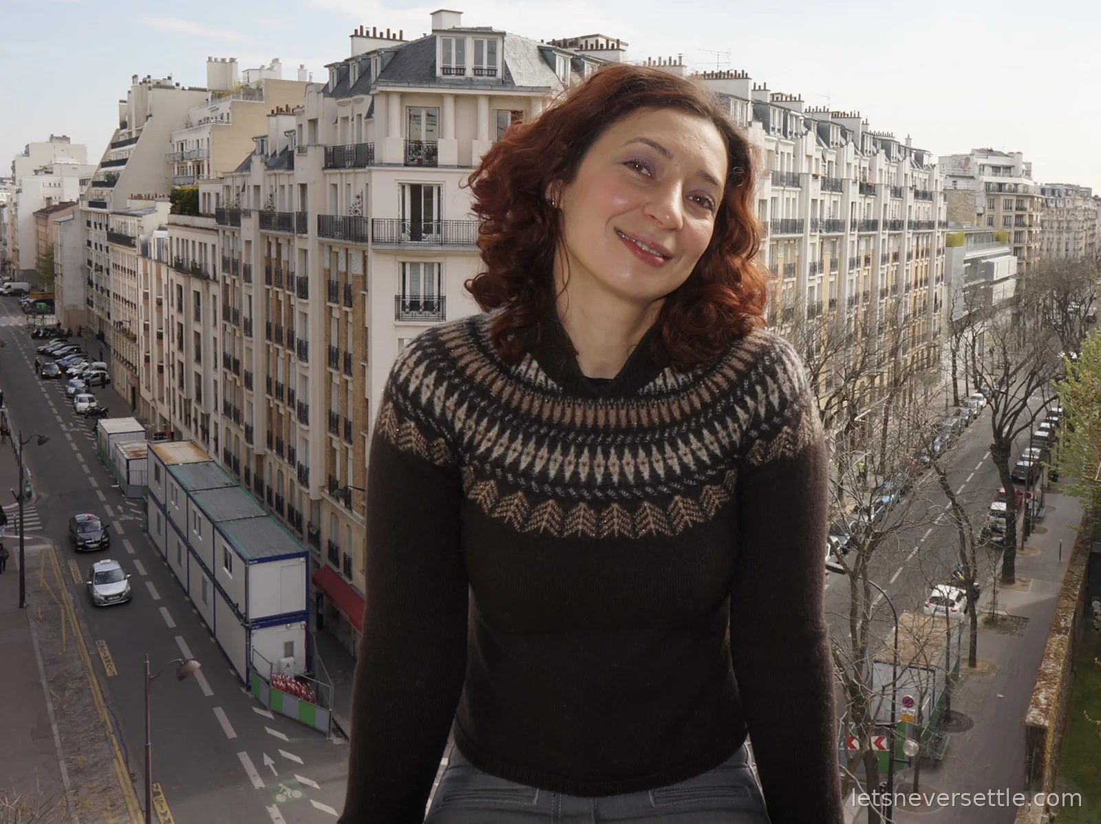 Inga on a high balcony above a Paris street lined with cream stone apartment buildings, mansard roofs and bare trees