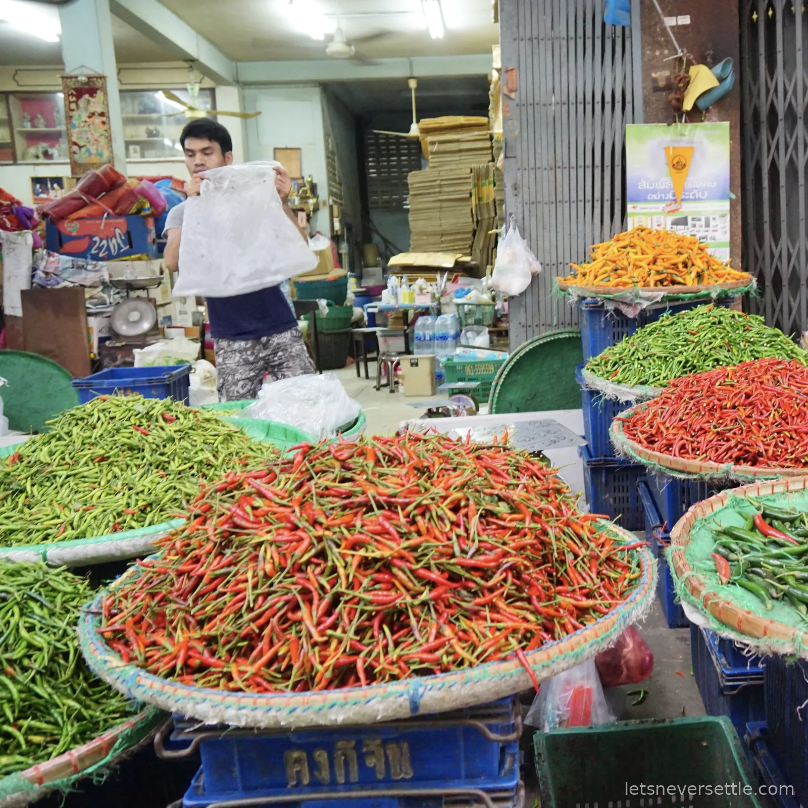 Wide flat woven baskets heaped with red, green and orange bird's eye chillies at a market stall, a vendor bagging up in the shop behind