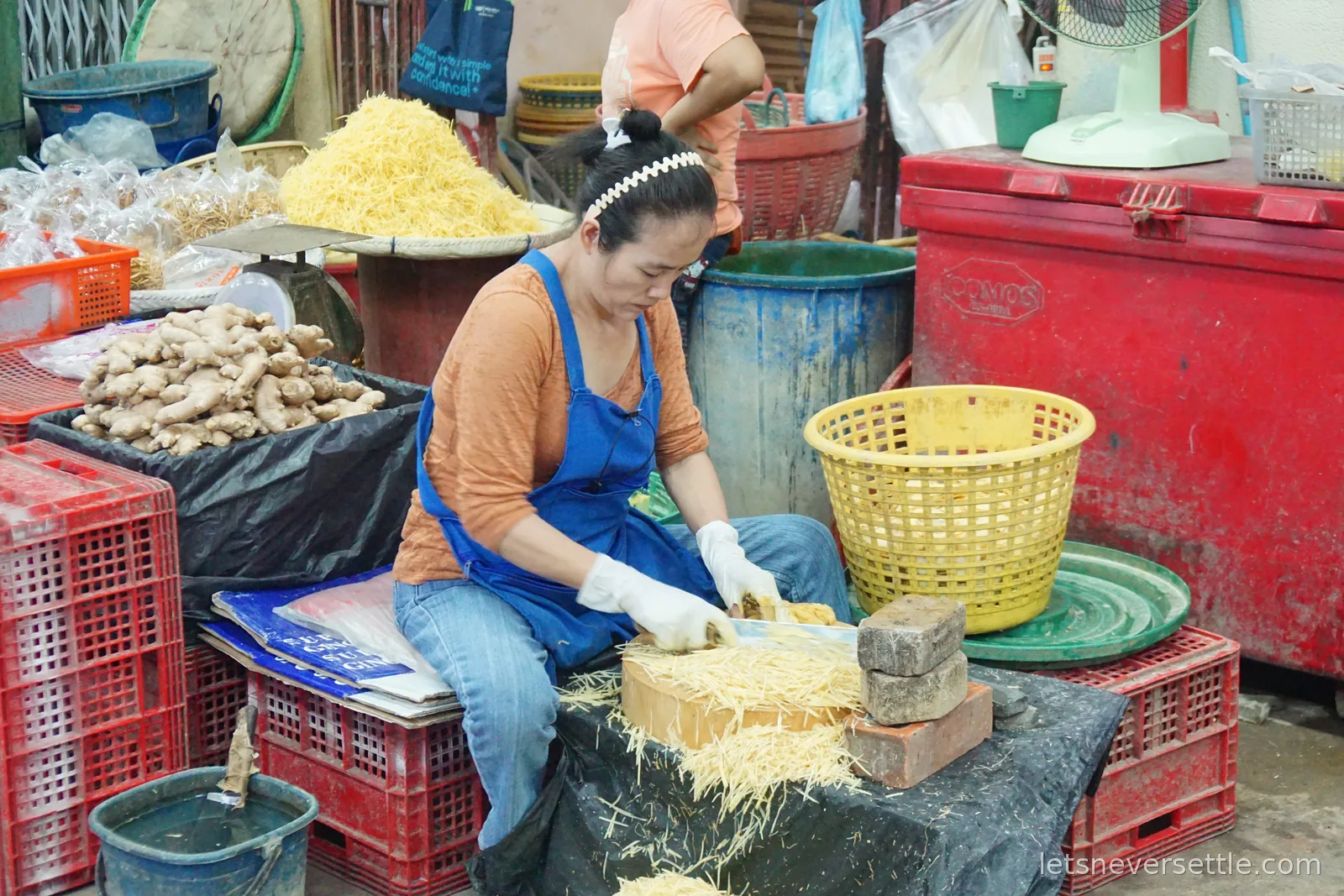 A woman in an orange top, blue apron and white gloves sitting on a crate shredding ginger into fine strips on a board weighted down with two bricks