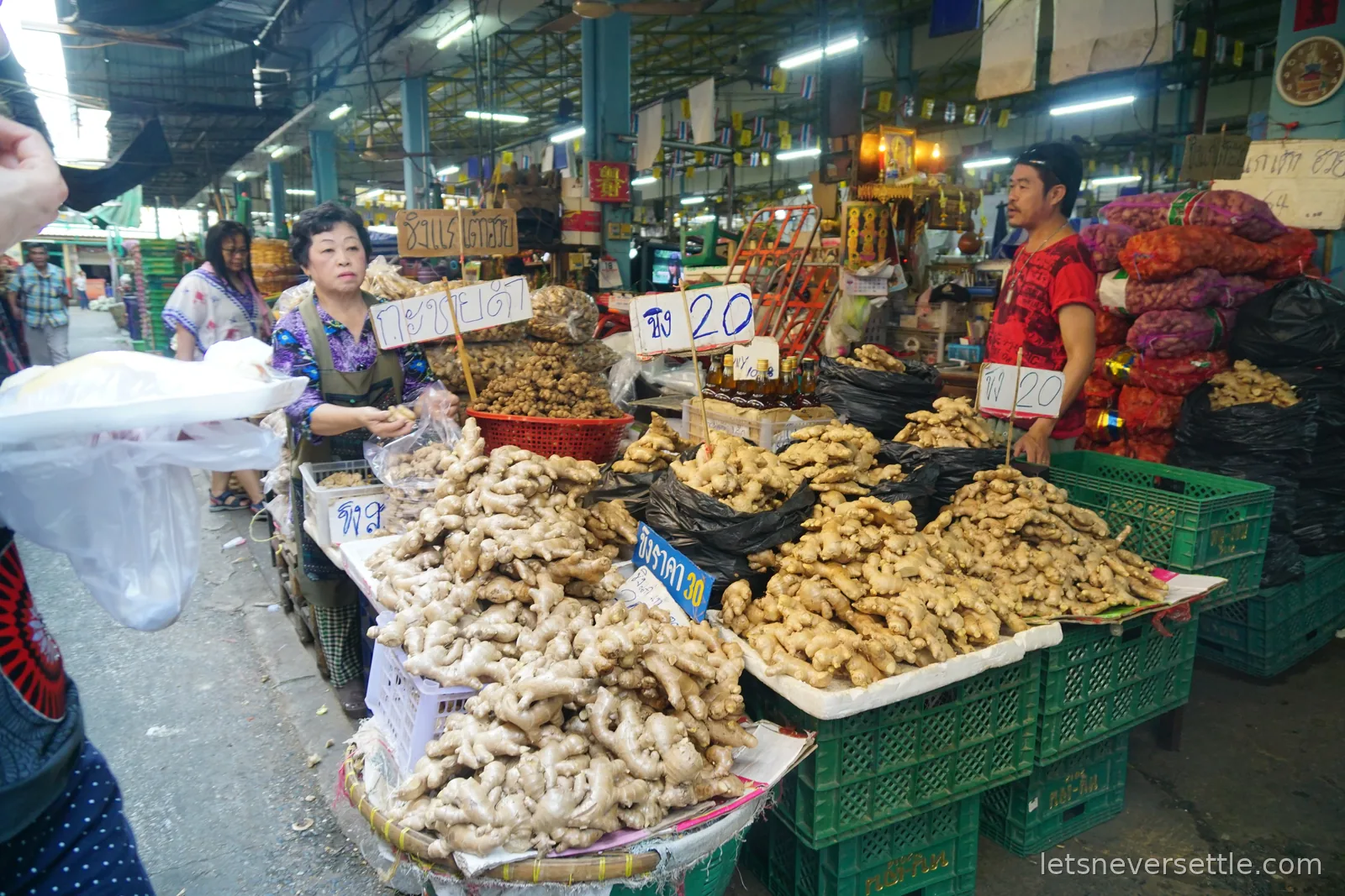 A ginger stall in a Thai market with trays of fresh ginger root and hand-written price cards reading 20 baht, two vendors standing behind