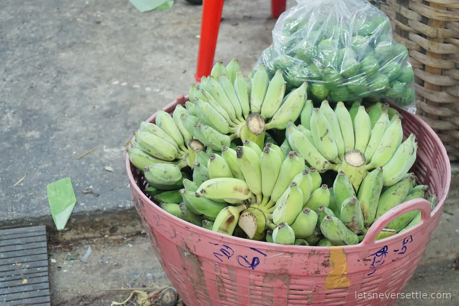 A pink plastic basket full of small green bananas on a market pavement, with a bag of green limes tucked in behind it