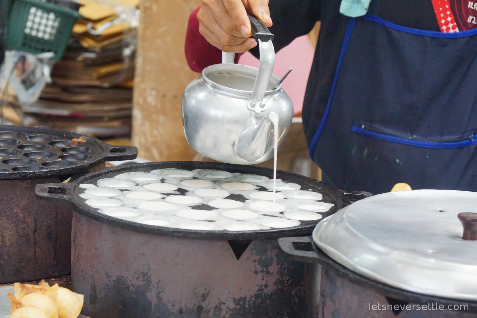 Close-up of a hand pouring white coconut batter from an aluminium kettle into the dimples of a hot cast iron pan