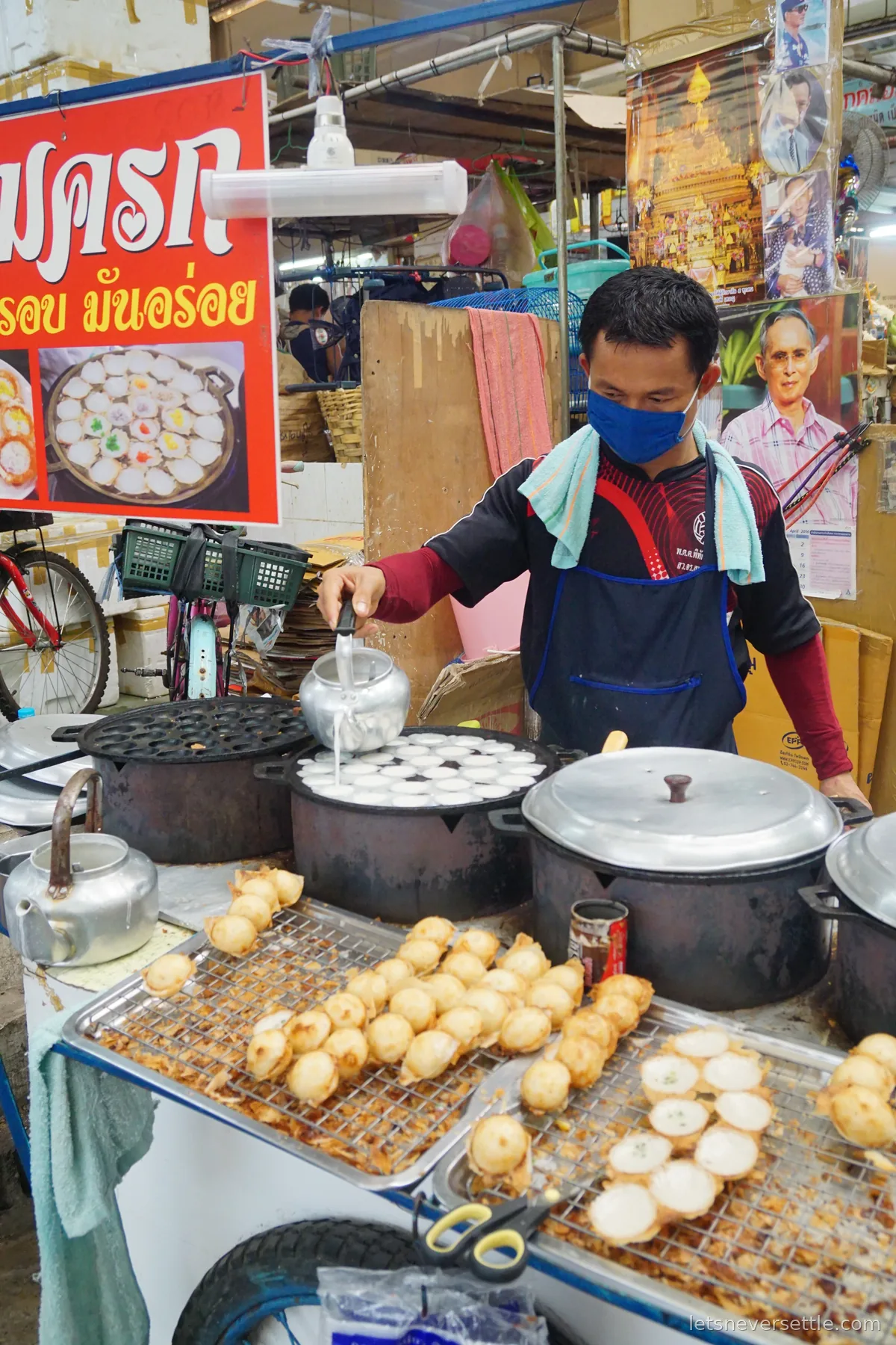 A vendor in a face mask and apron working a cart of dimpled cast iron pans making coconut pancakes, under a red sign showing a plate of the finished pancakes