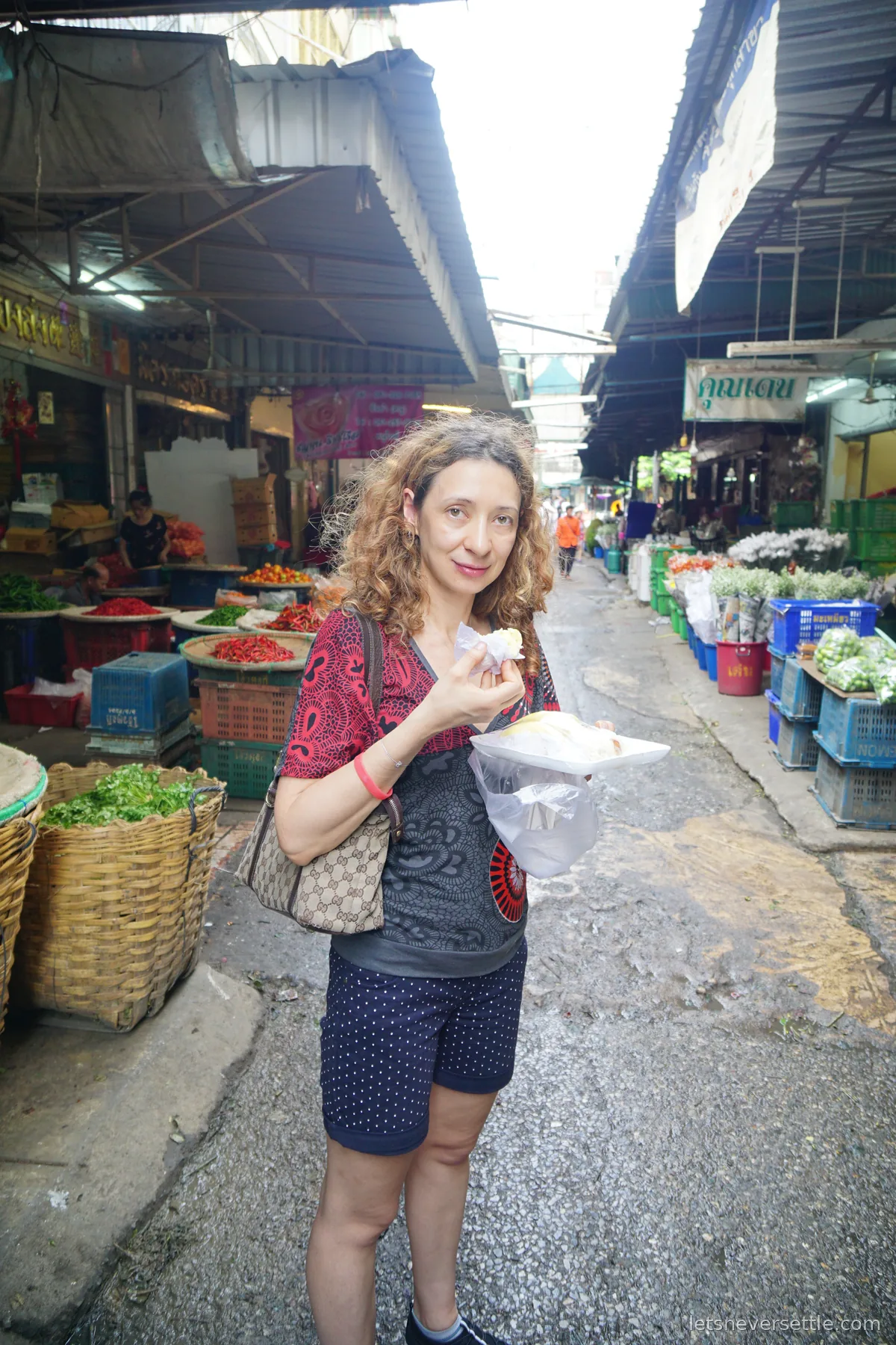 Inga eating a coconut pancake in a covered market lane, baskets of red and green chillies on one side and blue crates of cut flowers on the other