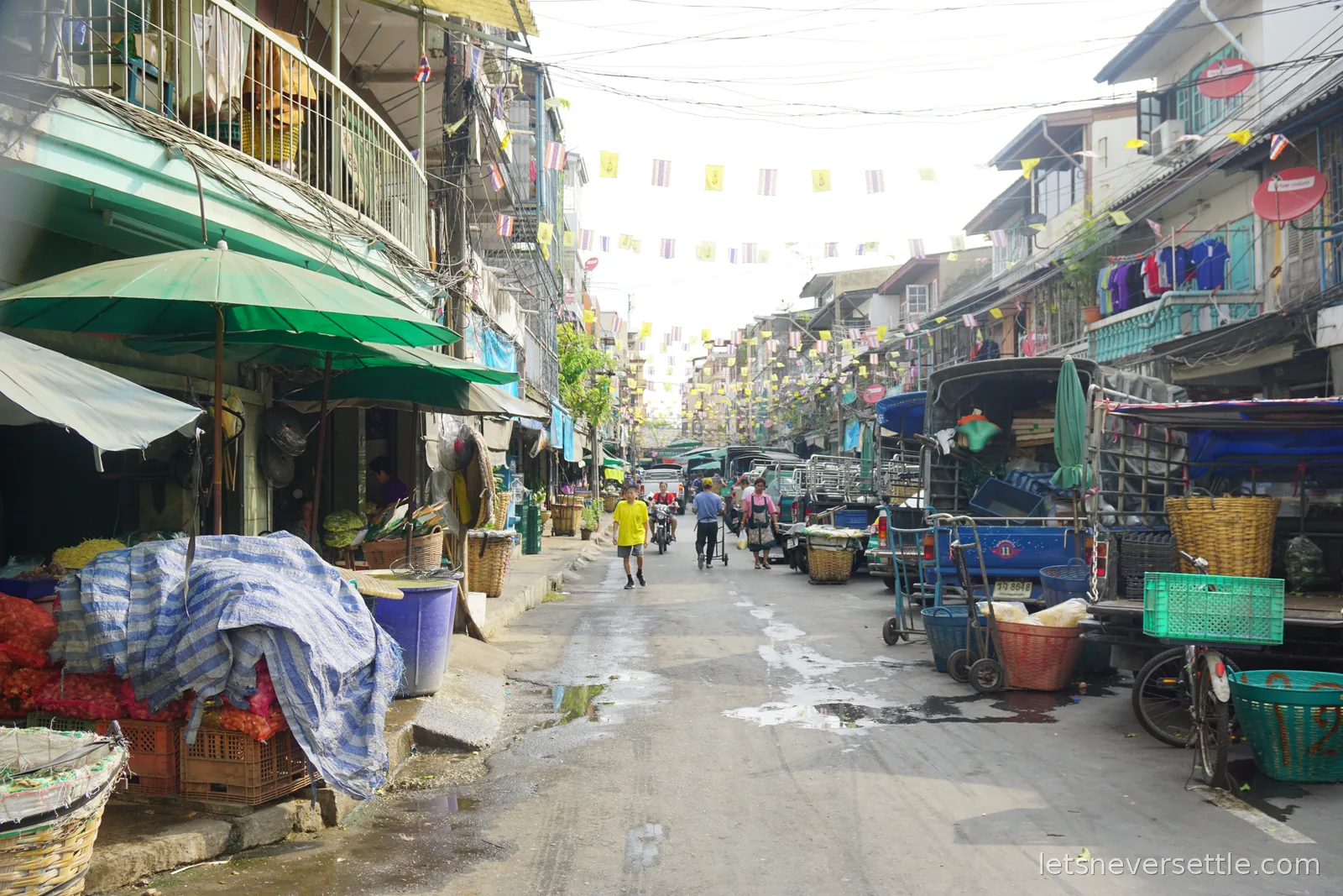 A market street lined with old shophouses, strung end to end with rows of yellow royal flags and Thai national flags, handcarts and pickup trucks parked along one side