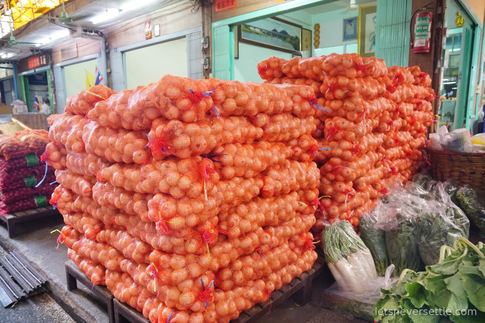 Pallets stacked high with orange mesh sacks of shallots and onions outside a green-shuttered shophouse in a Thai market