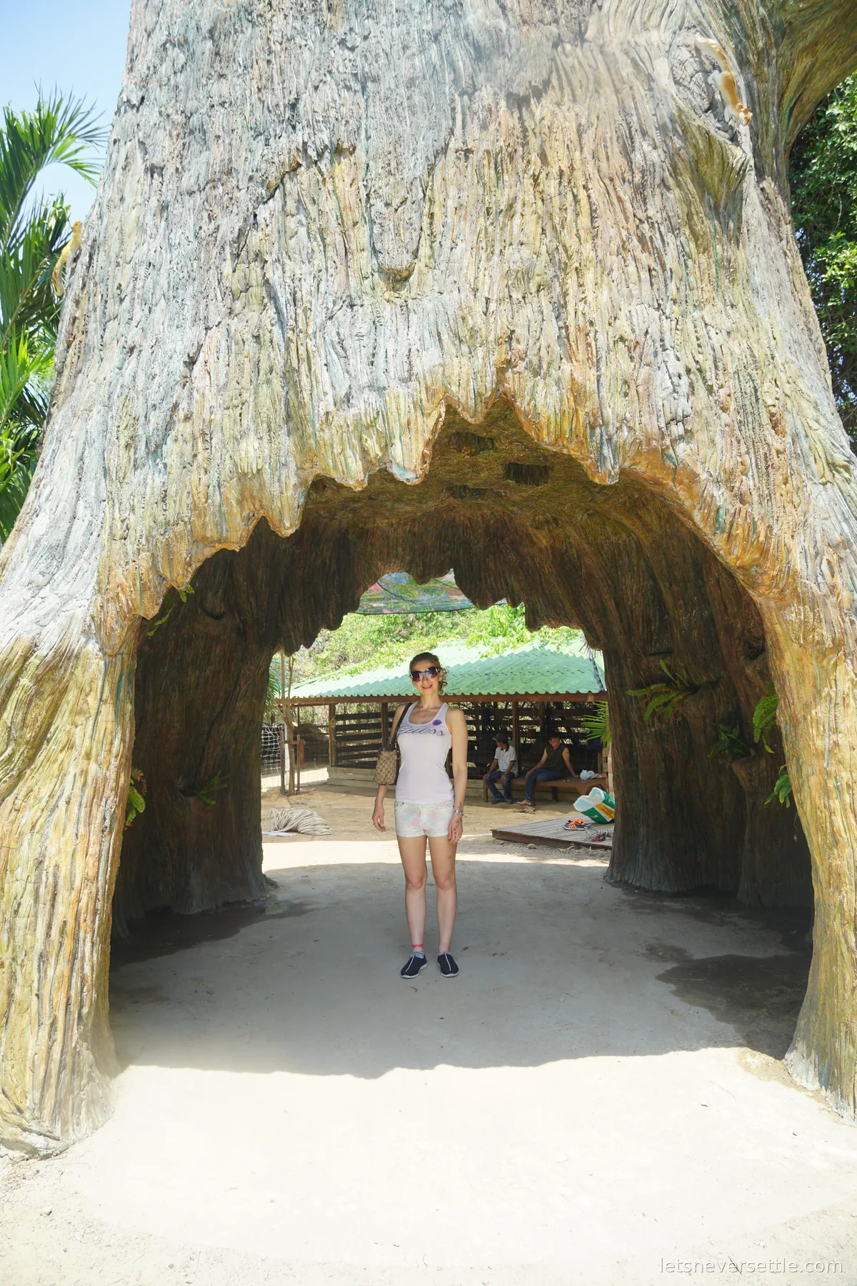 Inga standing inside an archway cut through the base of an enormous petrified tree trunk, a green-roofed shelter visible through the opening behind her