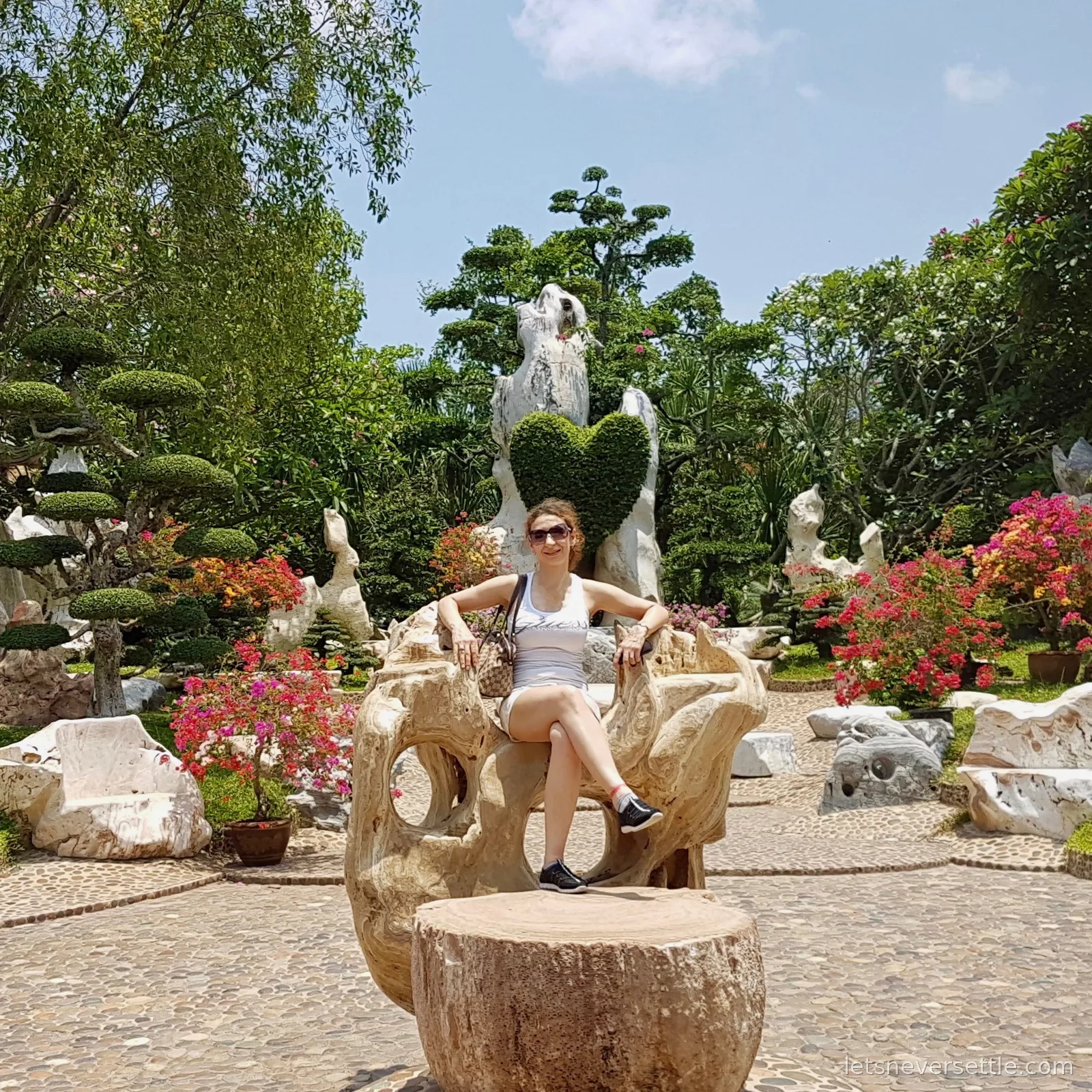 Inga sitting on a carved petrified wood bench in a garden of cloud-pruned topiary, pale stone sculptures and bright bougainvillea