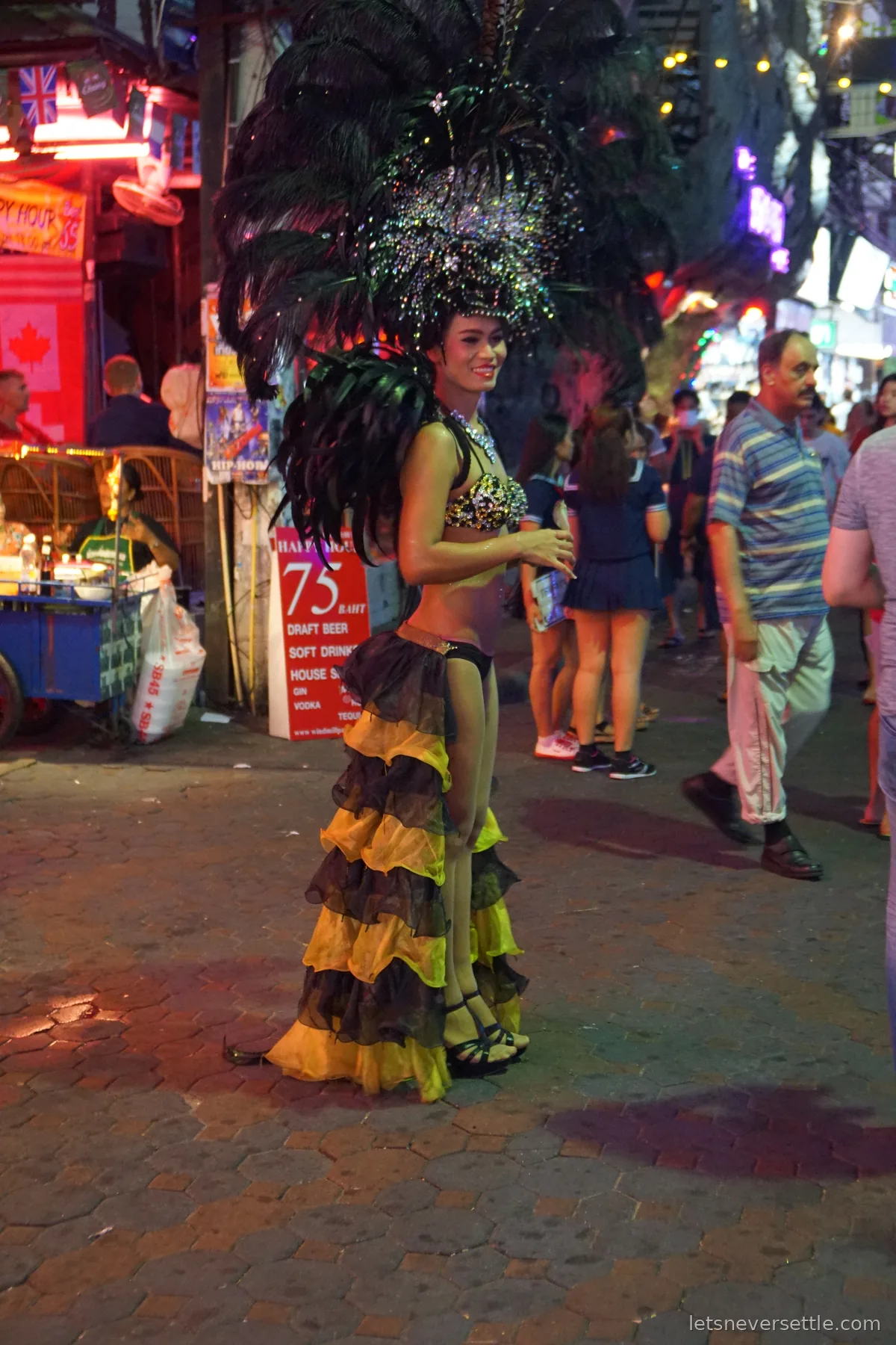 A cabaret performer in a sequinned costume and a tall black feather headdress standing on Pattaya's Walking Street at night, tourists walking past