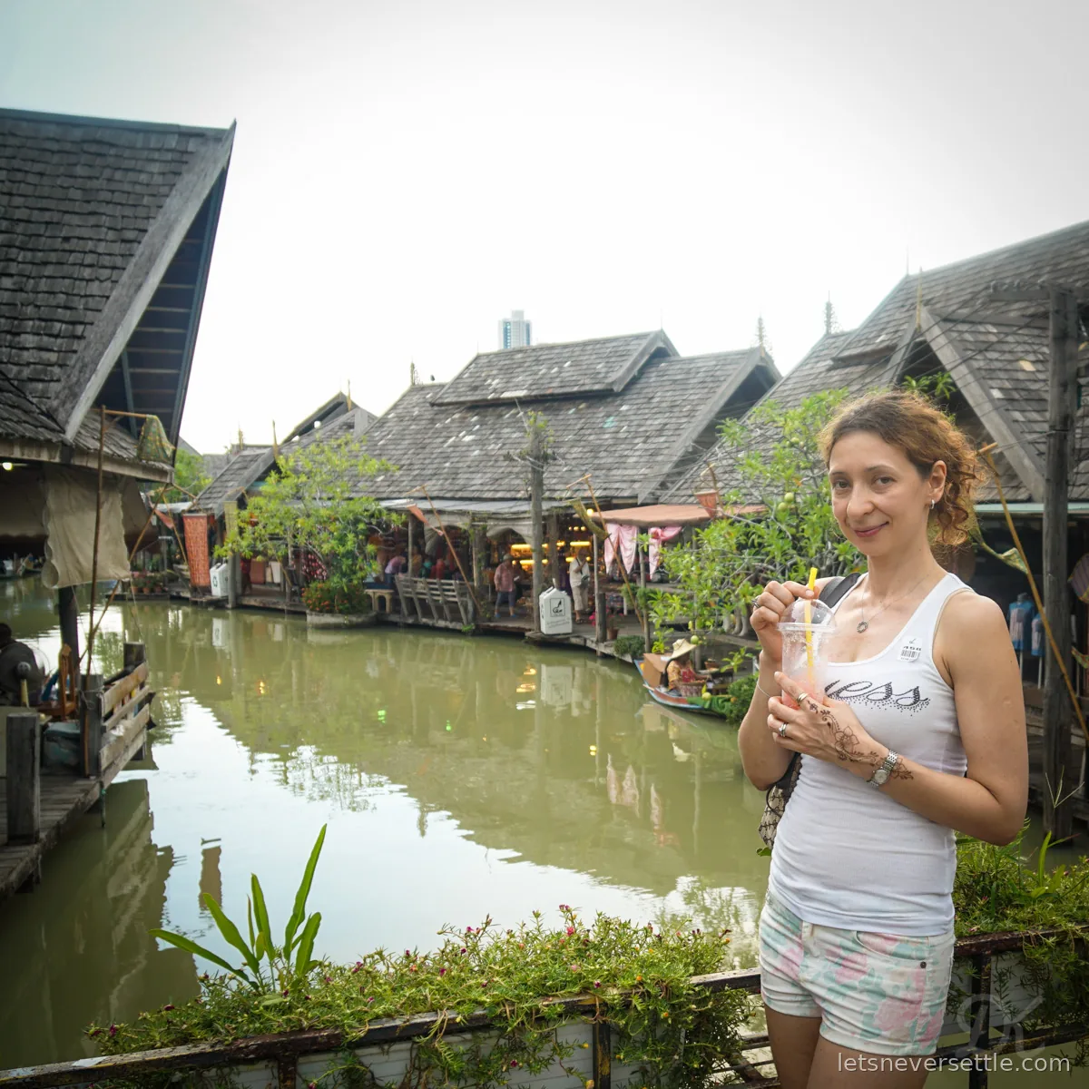 Inga holding an iced drink beside a canal at Pattaya's floating market, wooden shingle-roofed shops on stilts lining the water behind her