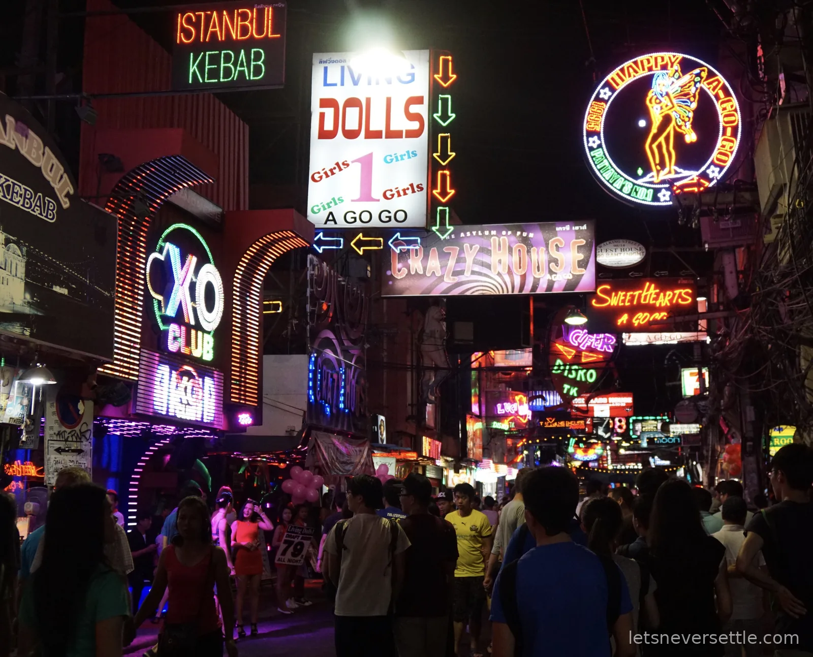 Crowds walking under a wall of neon signs on Pattaya's Walking Street at night, including a kebab shop, go-go bars and a club sign