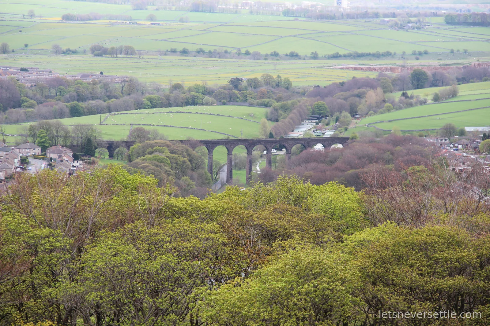 View from Grin Low over the treetops to a stone railway viaduct arching across a green valley of fields and dry stone walls