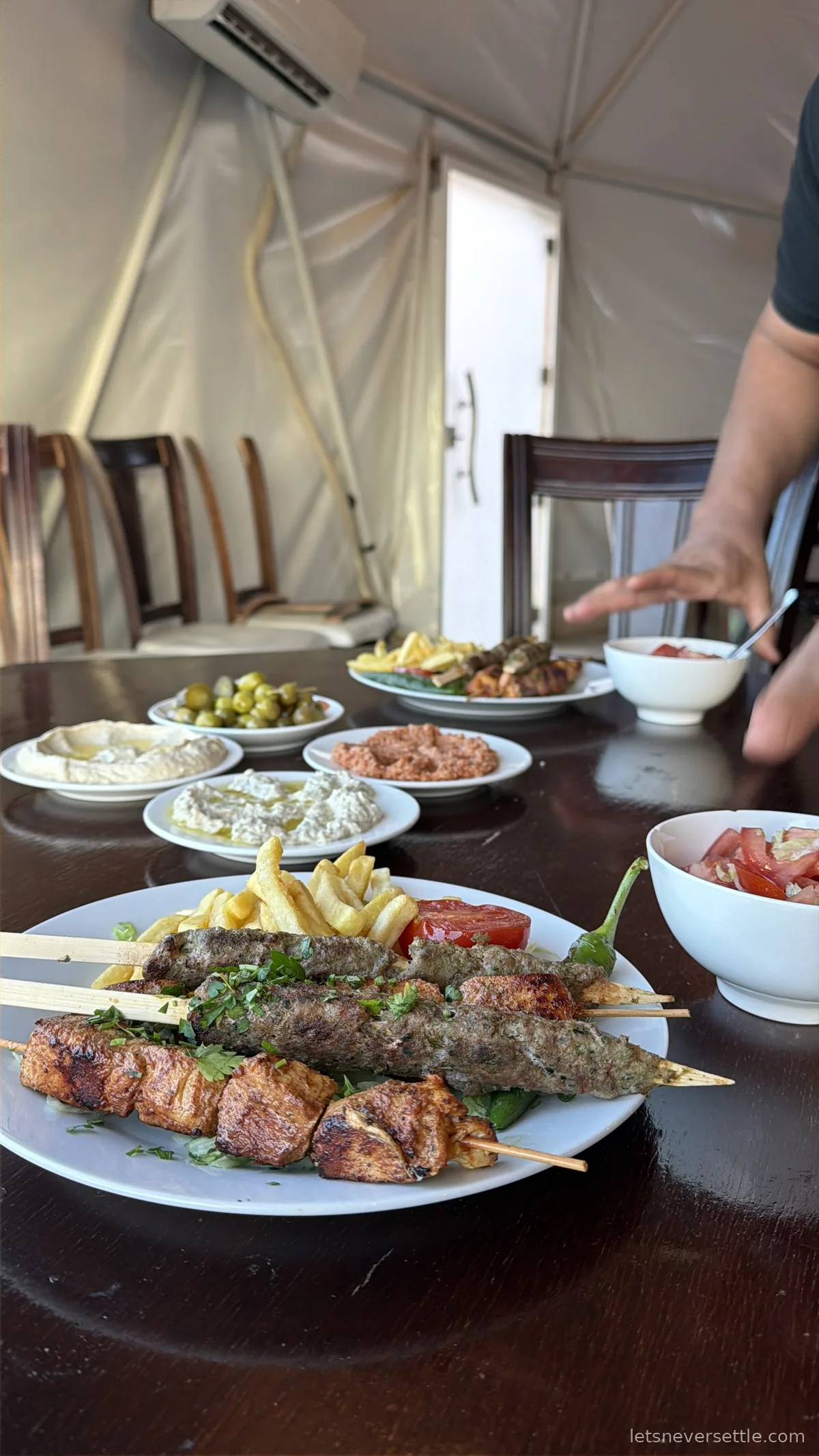 A plate of grilled kofta and chicken skewers with chips, beside bowls of hummus, olives and salads on a table in the camp restaurant
