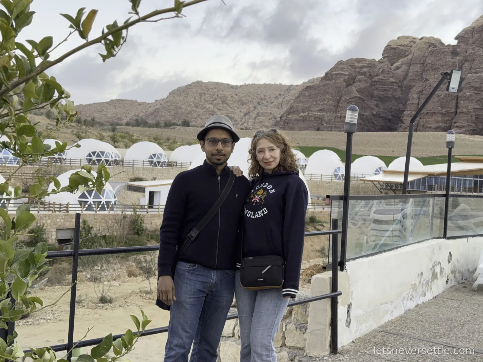 Inga and Dileep standing at a railing in front of the white dome tents of their camp near Petra, with red sandstone cliffs rising behind