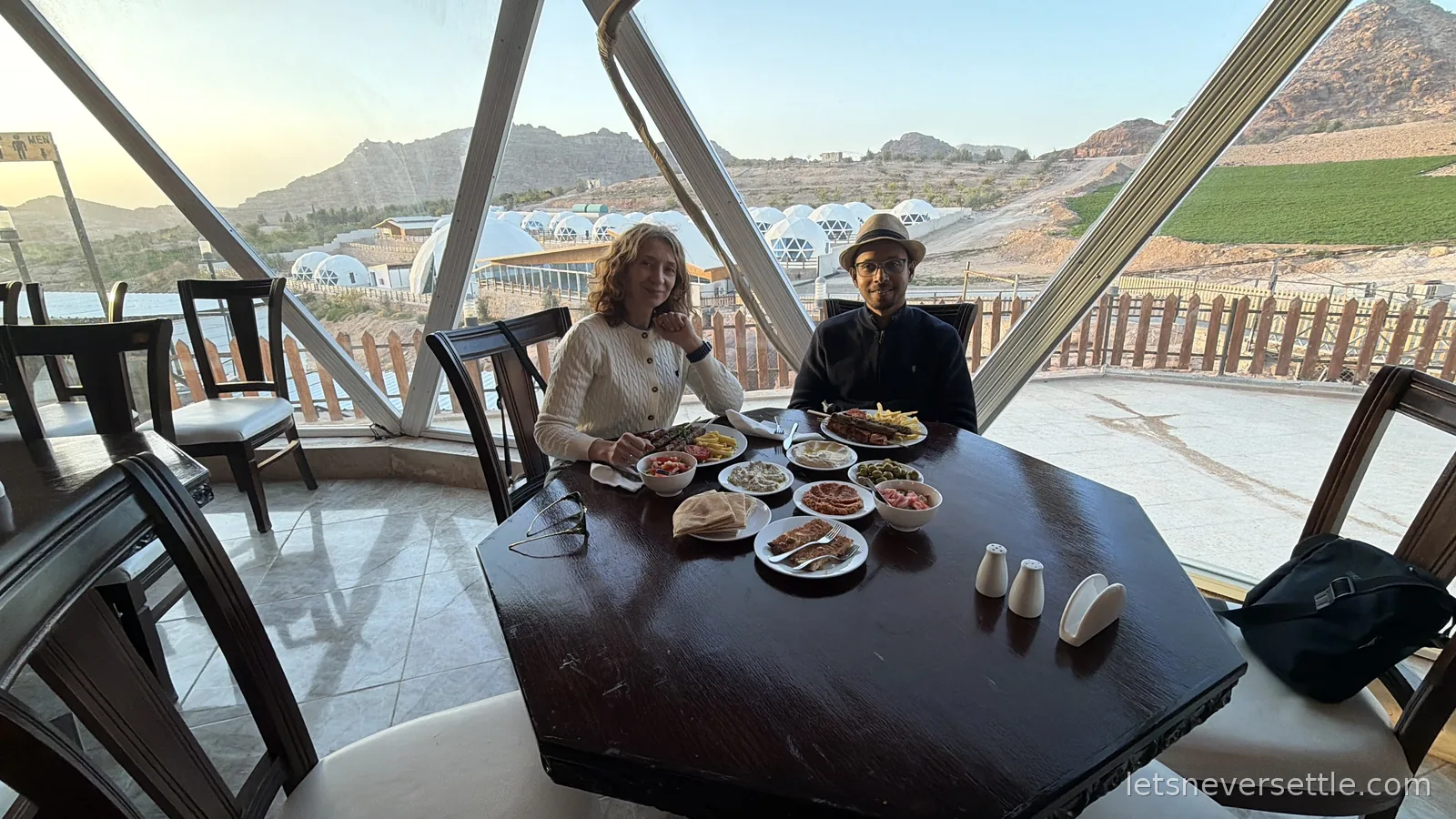 Inga and Dileep sitting at a table inside a glass-fronted dome restaurant, with the camp's white domes and the hills beyond visible through the window