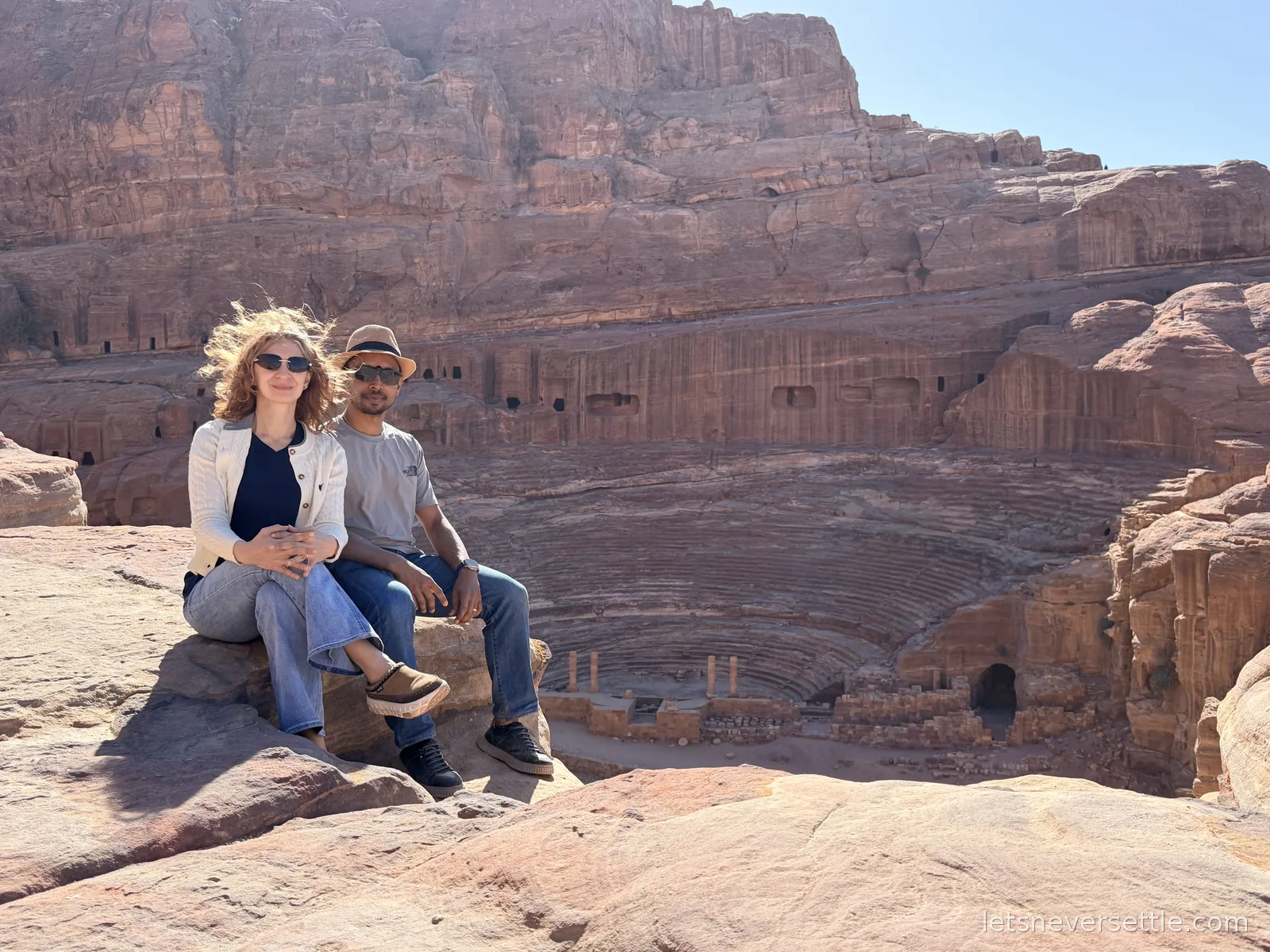 Inga and Dileep sitting on a rock above Petra's Roman Theatre, its curved rows of seating carved straight into the hillside behind them