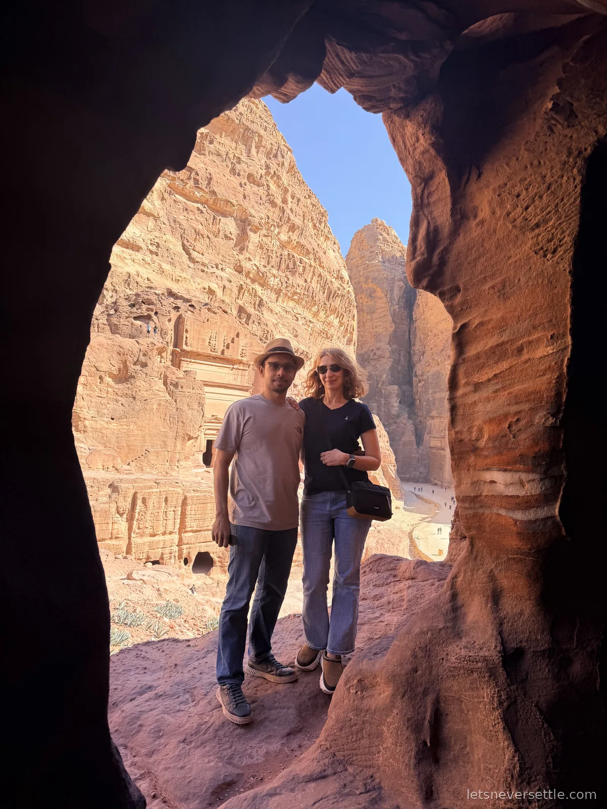 Inga and Dileep framed by the mouth of a rock cave at Petra, with the carved facade of the Royal Tombs on the cliff behind them