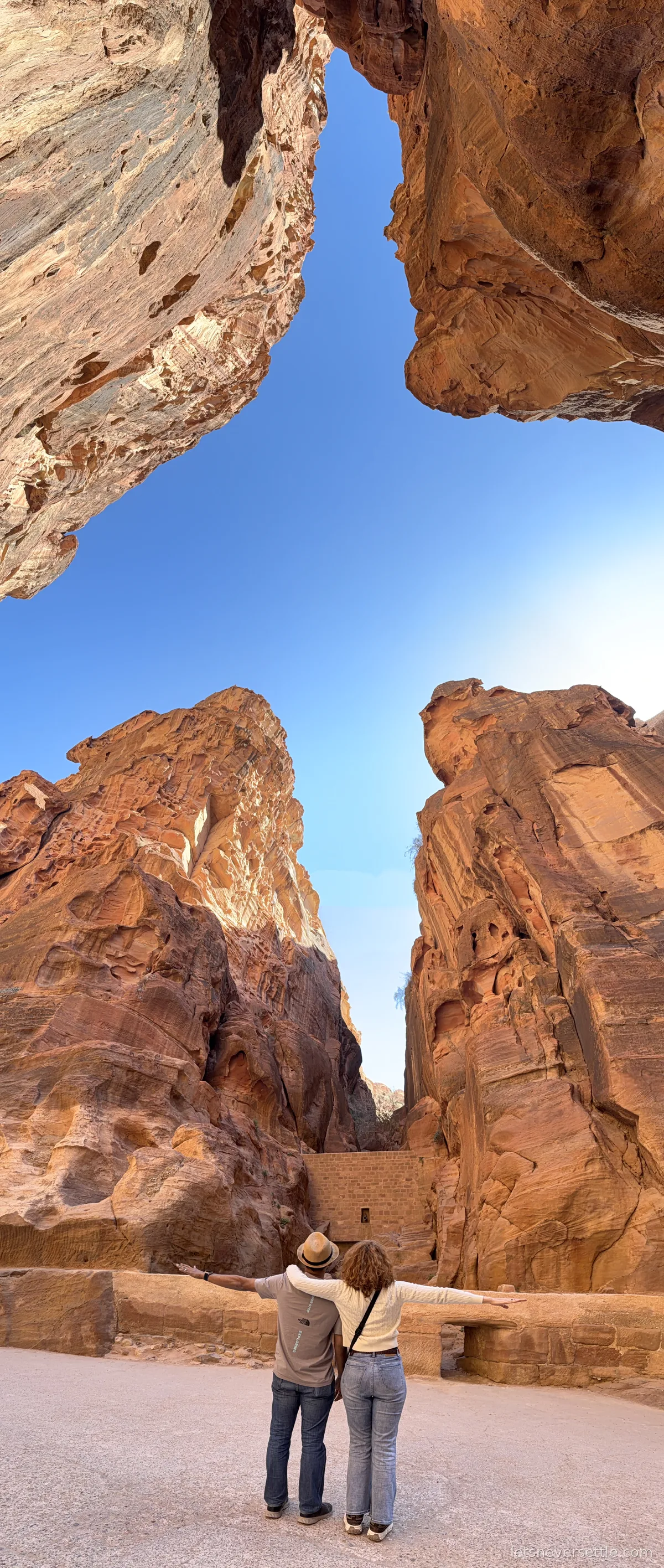 Inga and Dileep standing with their arms outstretched at the entrance to the Siq at Petra, huge overhanging sandstone boulders framing the strip of sky above them