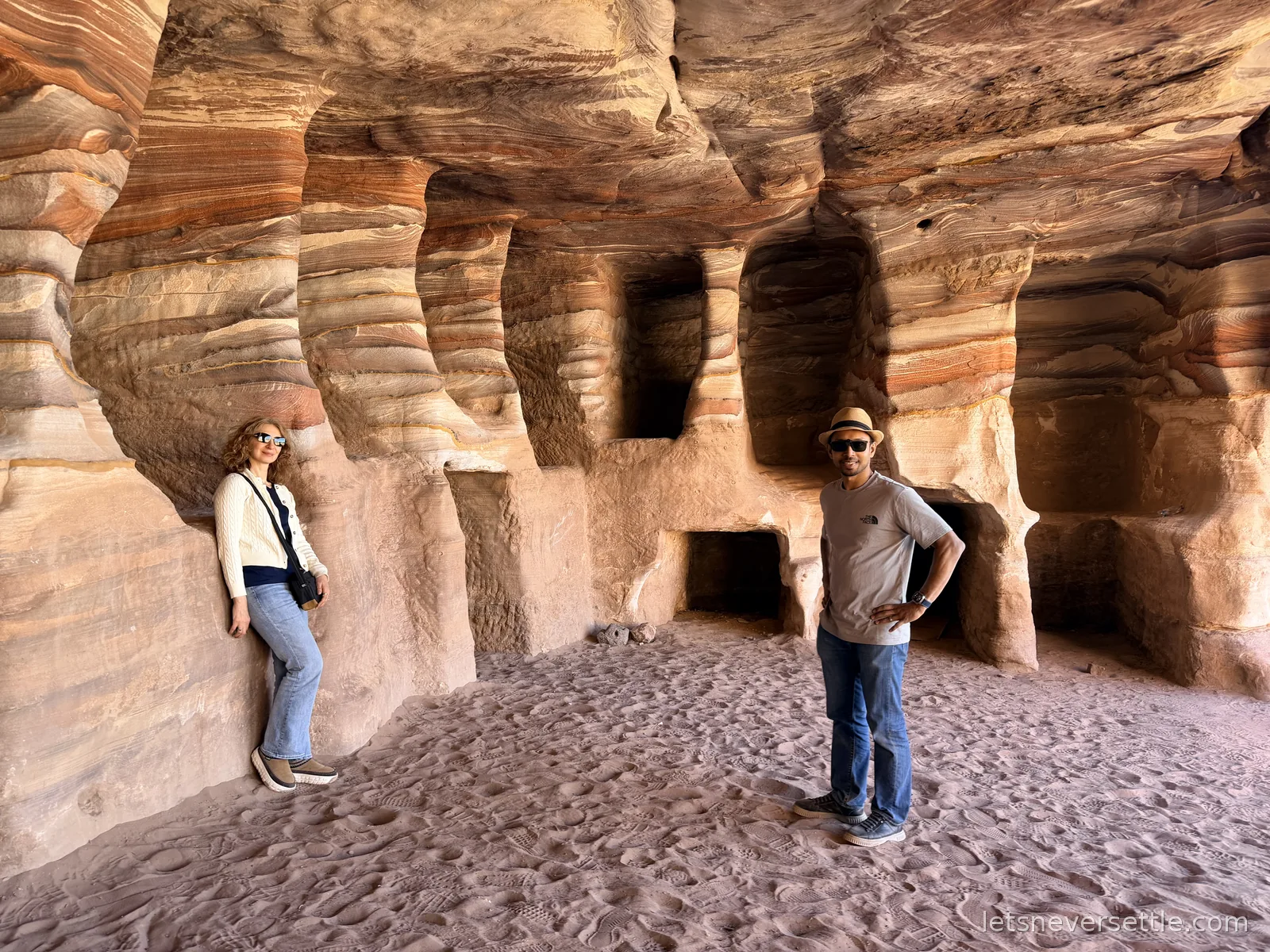 Inga and Dileep standing inside a large carved rock chamber at Petra, the walls swirling with red, orange and white bands of sandstone