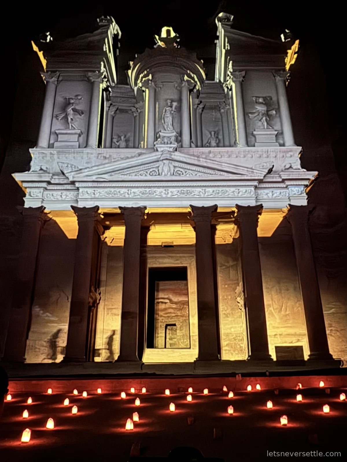 The Treasury facade at Petra lit in white and blue during the Petra by Night show, with rows of candles glowing red on the sand below