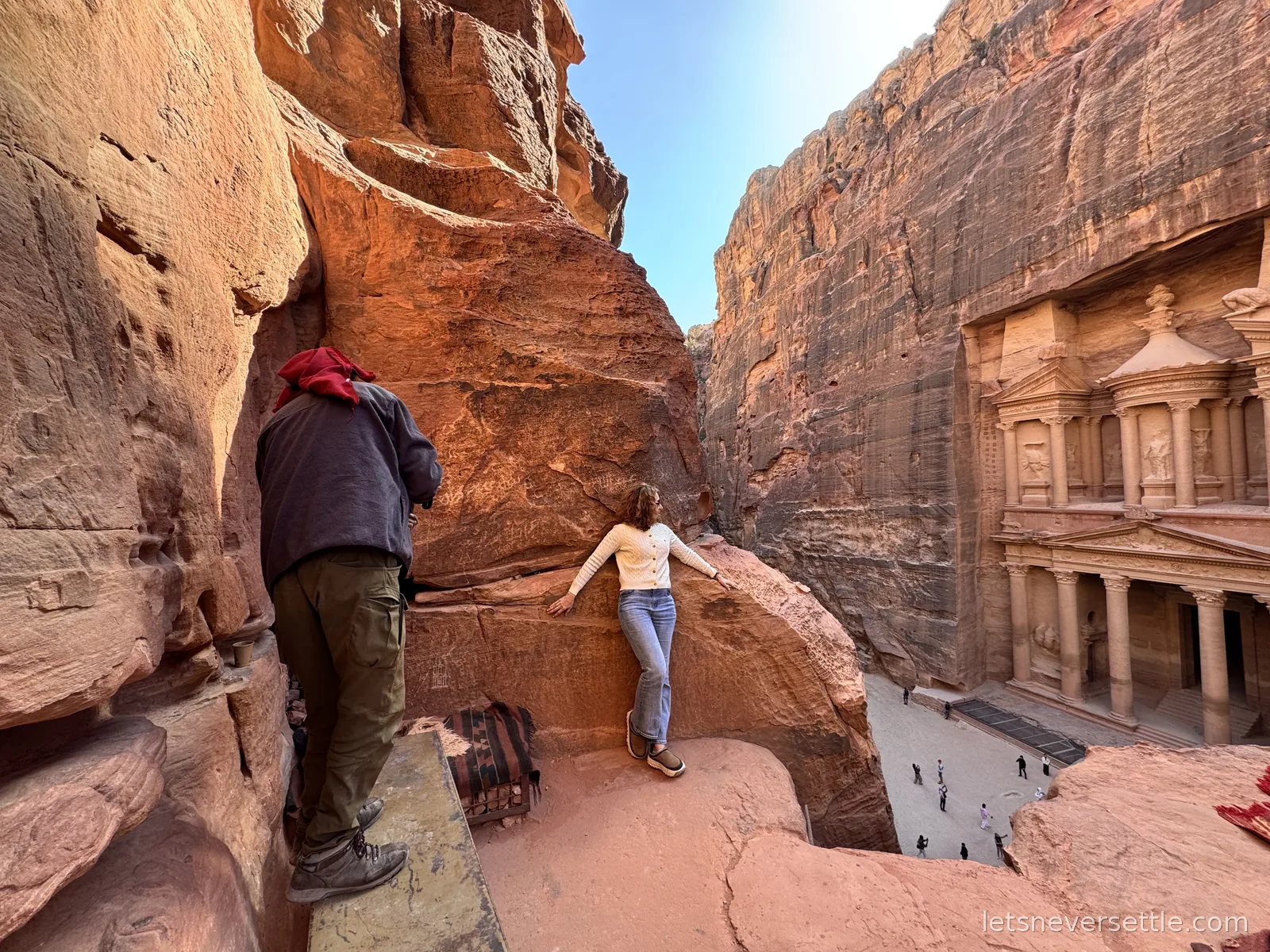 Inga climbing onto a rock ledge high above the Treasury at Petra, with a Bedouin guide ahead of her on the path and the Treasury facade in the canyon below