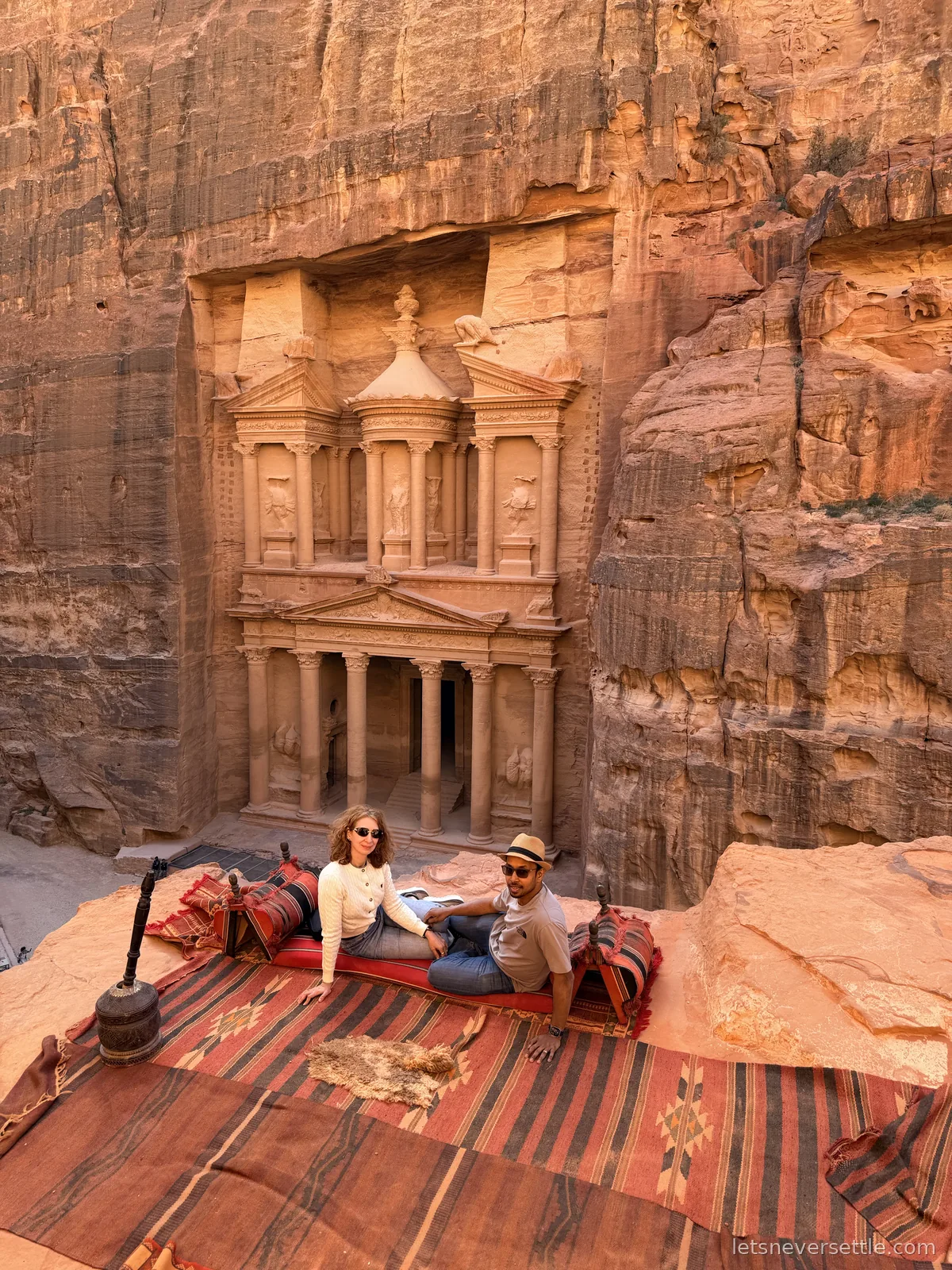 Inga and Dileep lounging on striped Bedouin rugs and cushions at a clifftop viewpoint above the Treasury at Petra