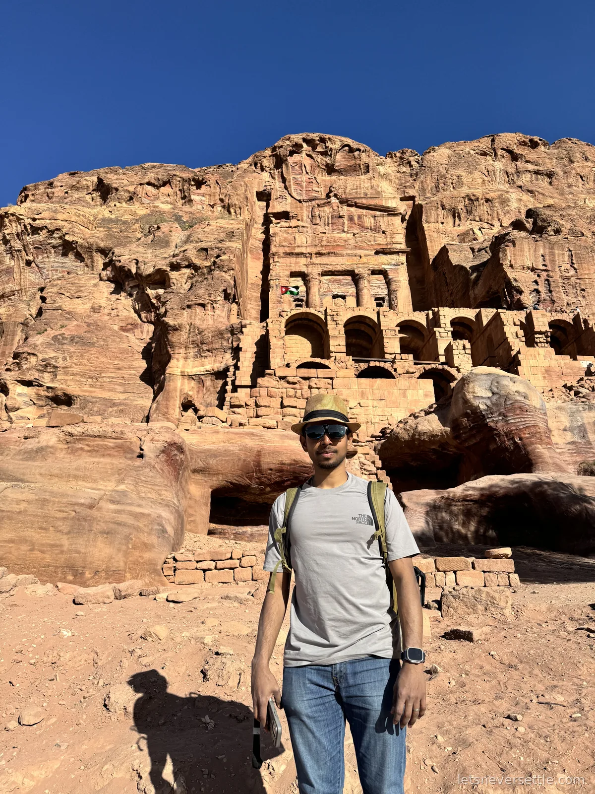Dileep standing below the Urn Tomb at Petra, its arched terrace and carved facade cut high into the red cliff behind him