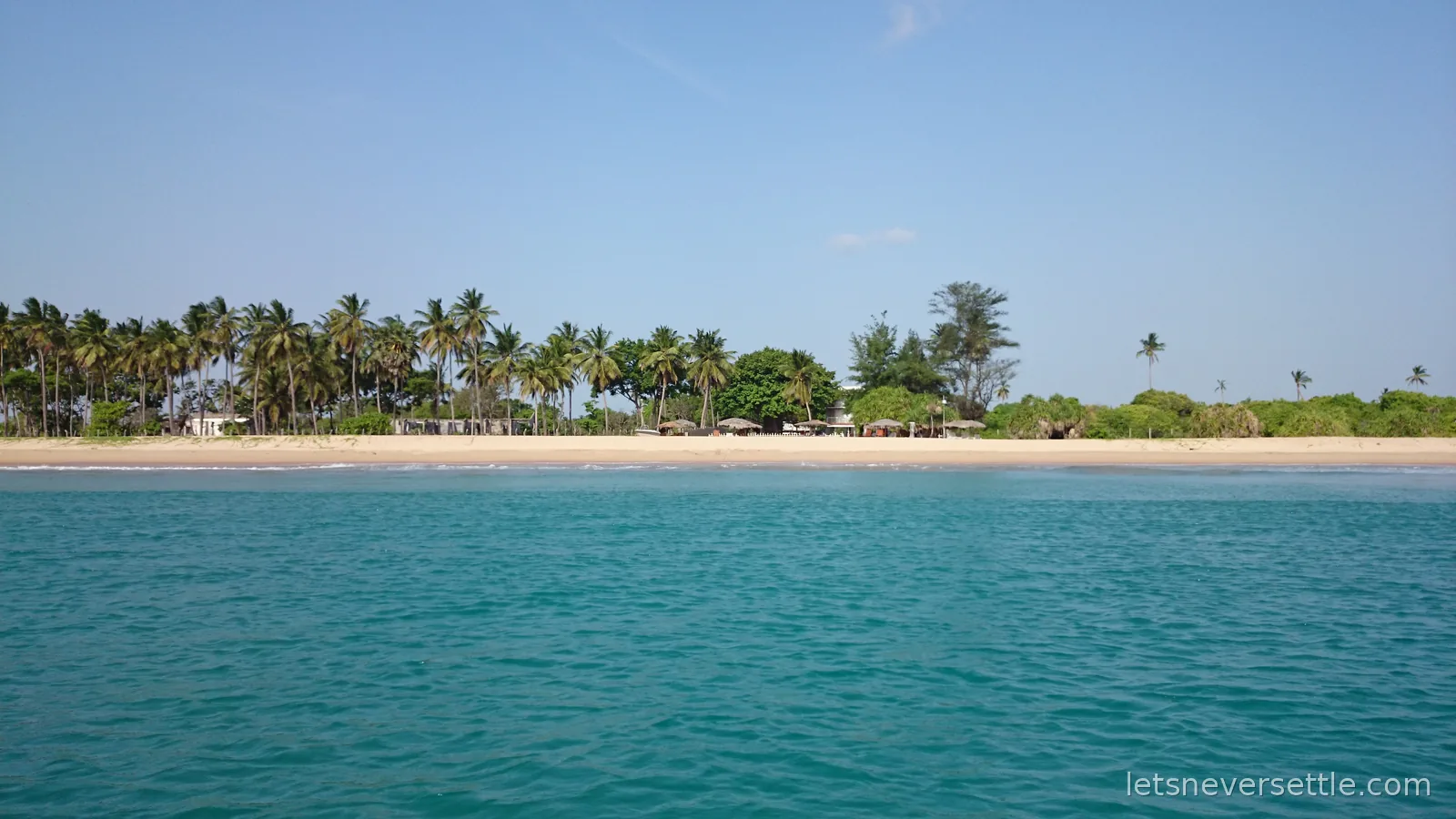 Nilaveli beach seen from the sea, a long strip of pale sand backed by coconut palms under a blue sky