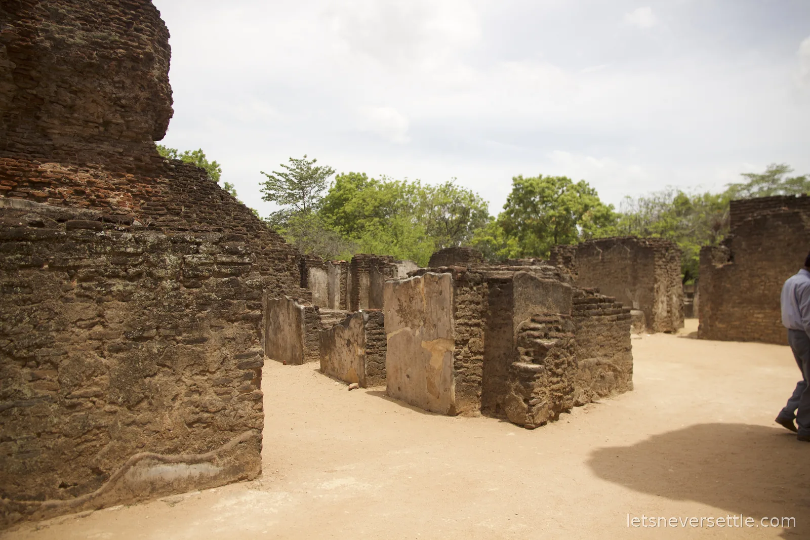 Weathered brick walls and plastered wall fragments of ruined buildings at Polonnaruwa, with sandy paths and green trees behind