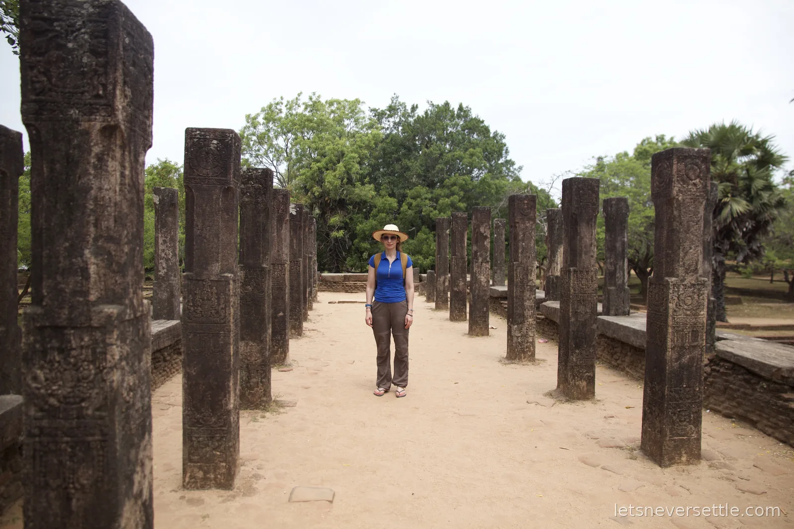 Inga in a sun hat and blue top standing on a sandy path between two rows of tall carved stone pillars at Polonnaruwa