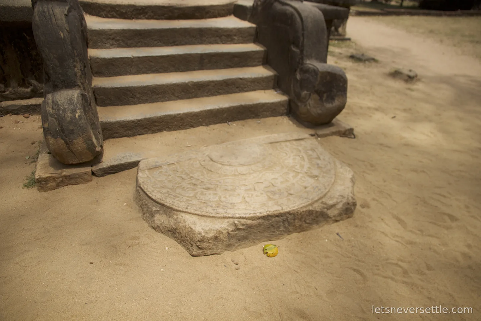 Seen through a stone doorway, an elderly woman in red bends to lay flowers at a small shrine inside a ruined stone temple at Polonnaruwa