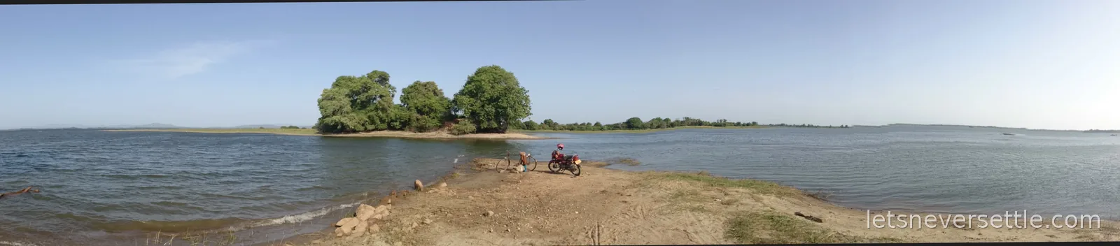 Panorama of Parakrama Samudra at Polonnaruwa: calm water on both sides of a sandy spit with an island of trees and a parked motorbike