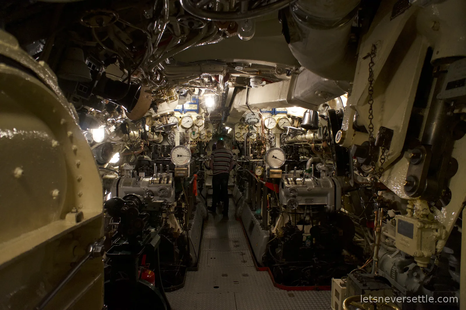 The narrow engine room of HMS Alliance, banks of machinery and dials either side of a walkway with a visitor at the far end