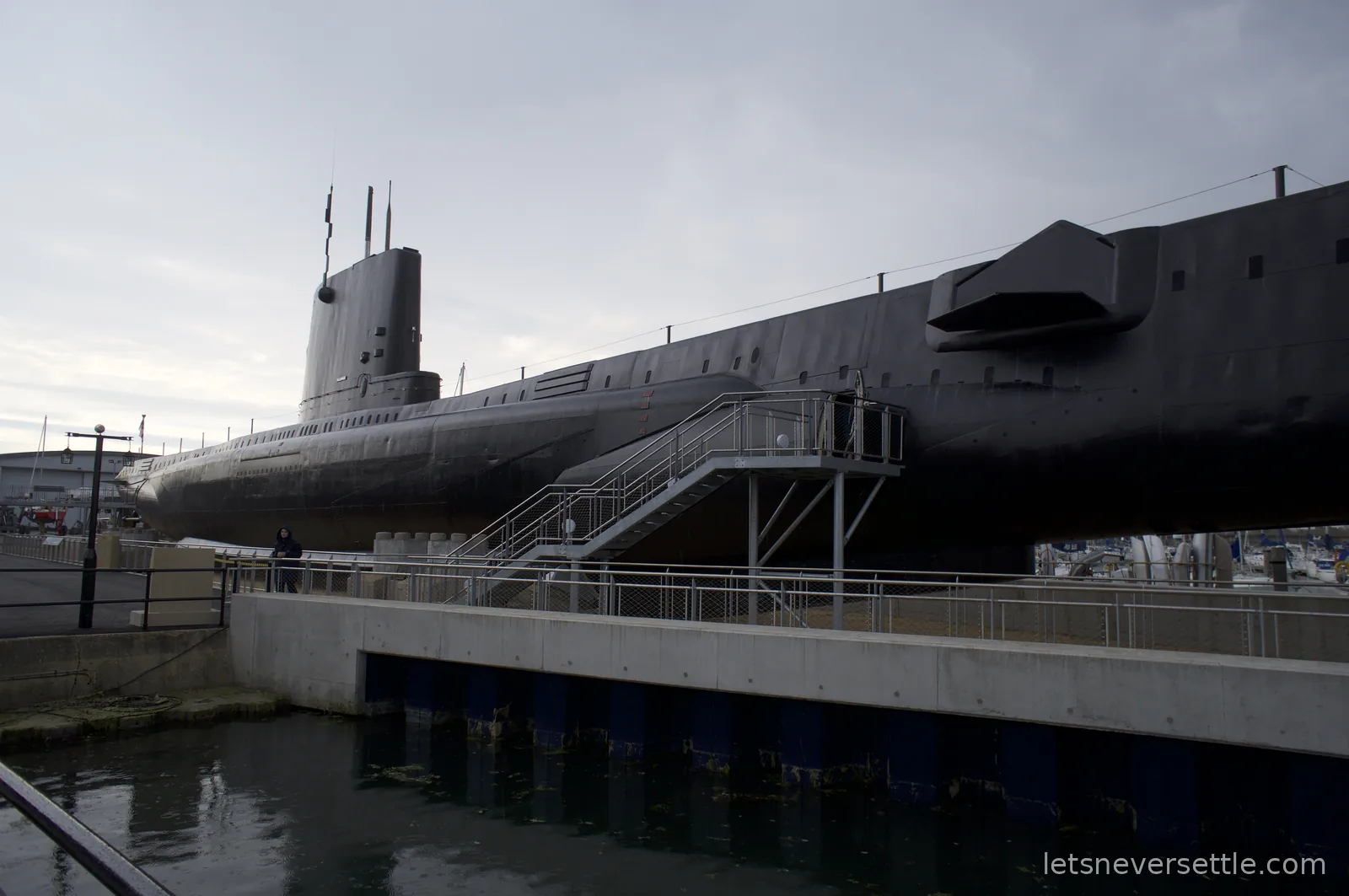 The long dark hull of the submarine HMS Alliance raised on supports beside the water at the Royal Navy Submarine Museum in Gosport, with an access staircase leading aboard