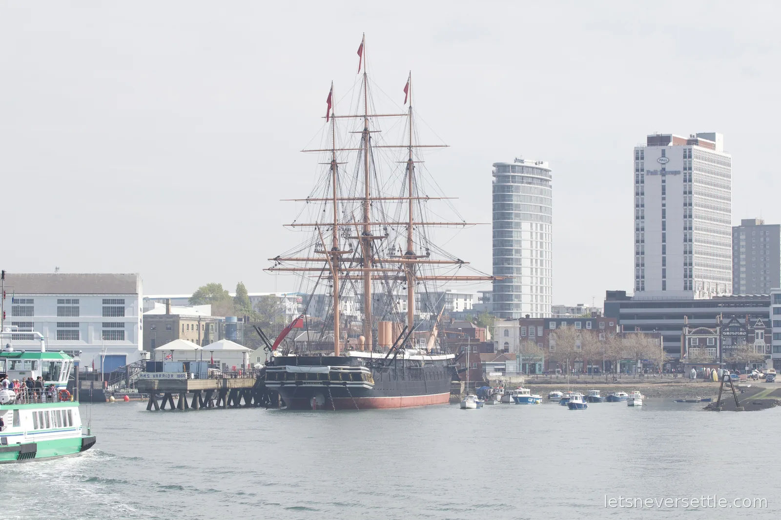 The black-hulled three-masted warship HMS Warrior moored at her jetty in Portsmouth harbour, modern tower blocks behind
