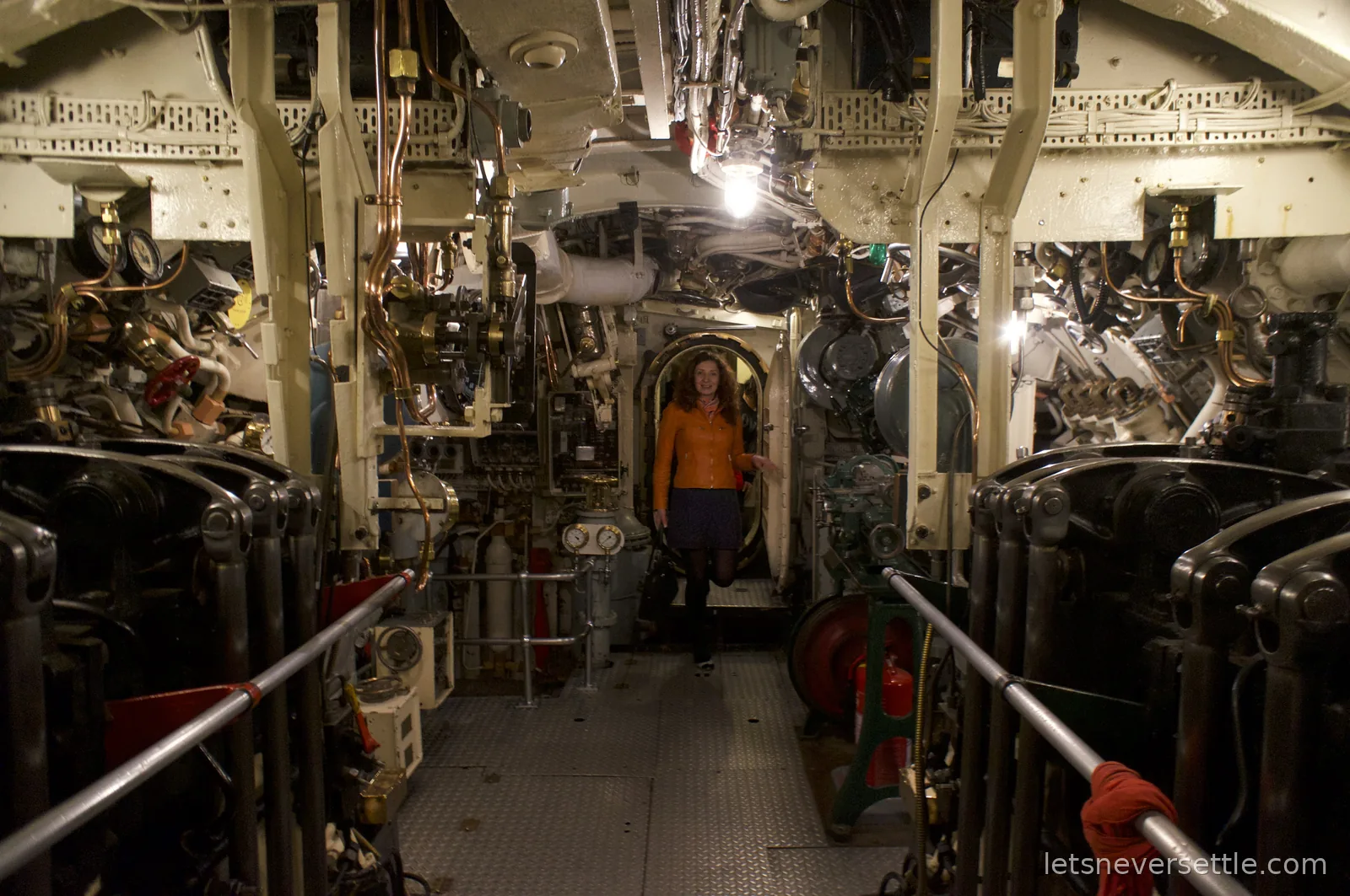Inga in an orange jacket standing at a hatch doorway in the machinery-lined interior of the submarine HMS Alliance