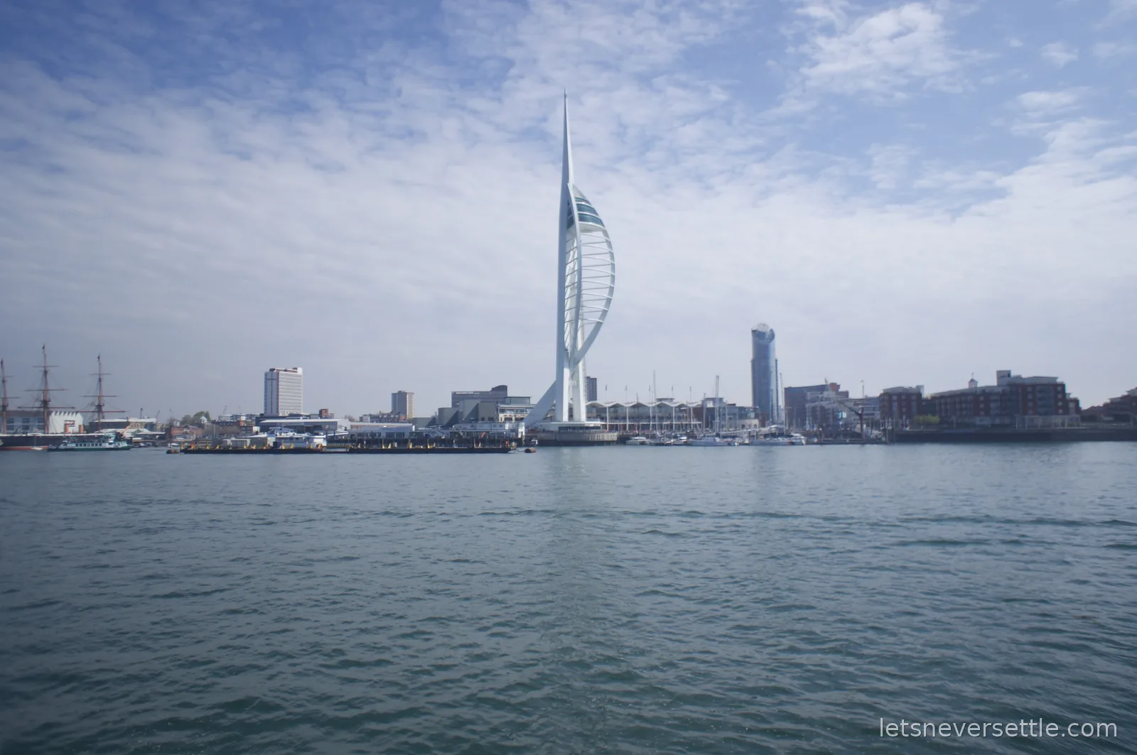 The white sail-shaped Spinnaker Tower rising over the Portsmouth waterfront, seen from out on the harbour with masts of historic ships to the left