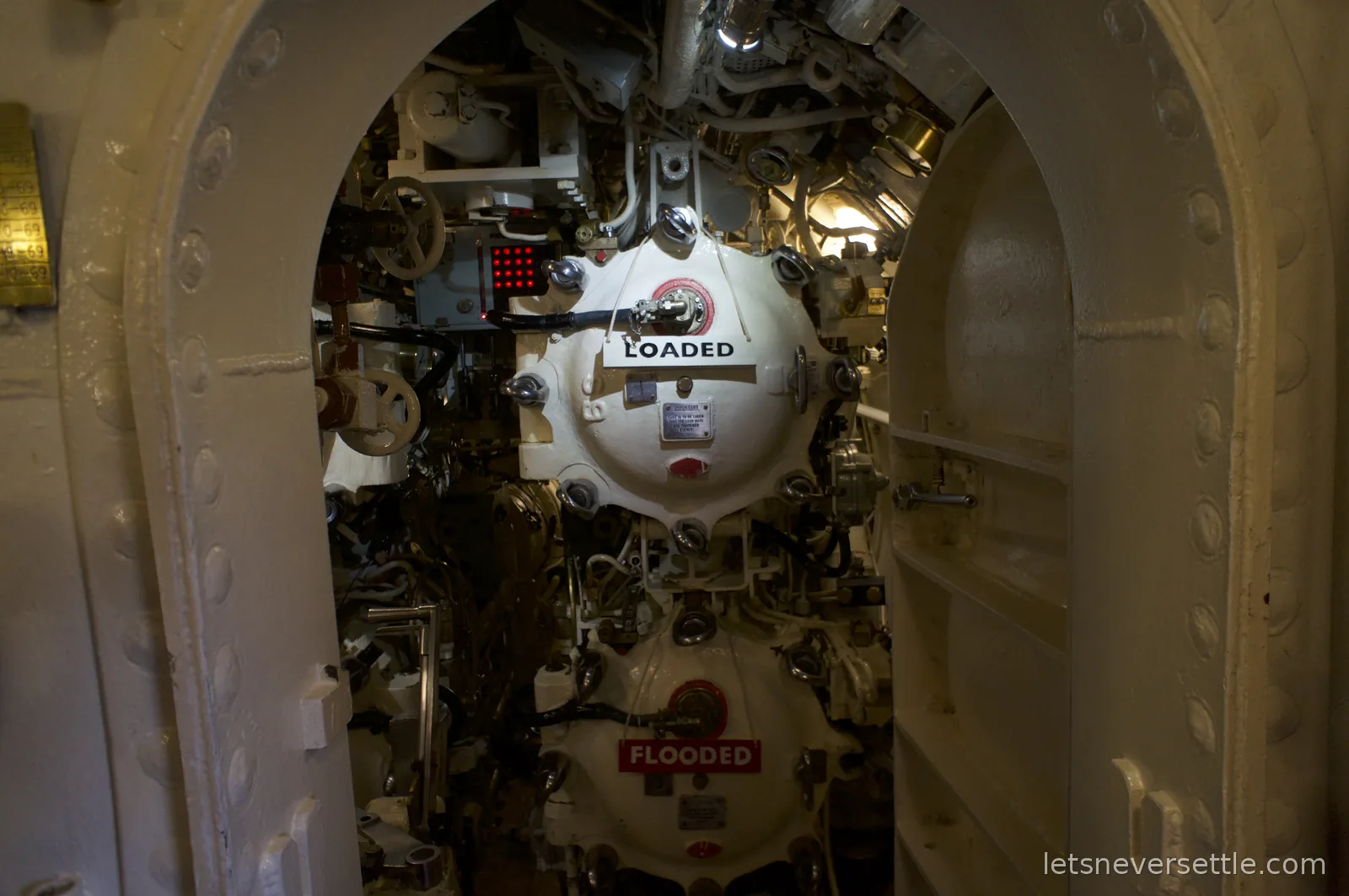 Torpedo tubes inside the submarine HMS Alliance seen through an oval hatchway, one tube labelled LOADED and another labelled FLOODED