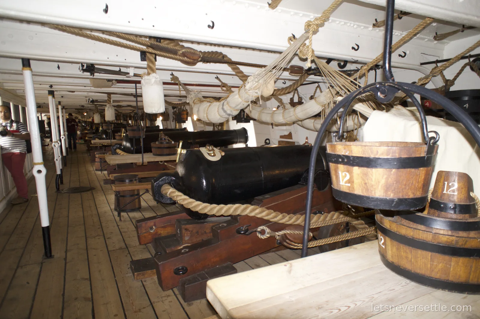 A gun deck aboard HMS Victory with a row of black cannon on wooden carriages, hammocks and rigging overhead and numbered wooden mess buckets hanging in the foreground