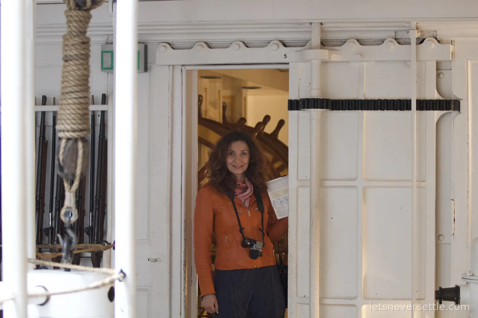 Inga in an orange jacket with a camera and guide leaflet standing in a white panelled doorway below decks, a large double ship's wheel and racks of rifles behind her