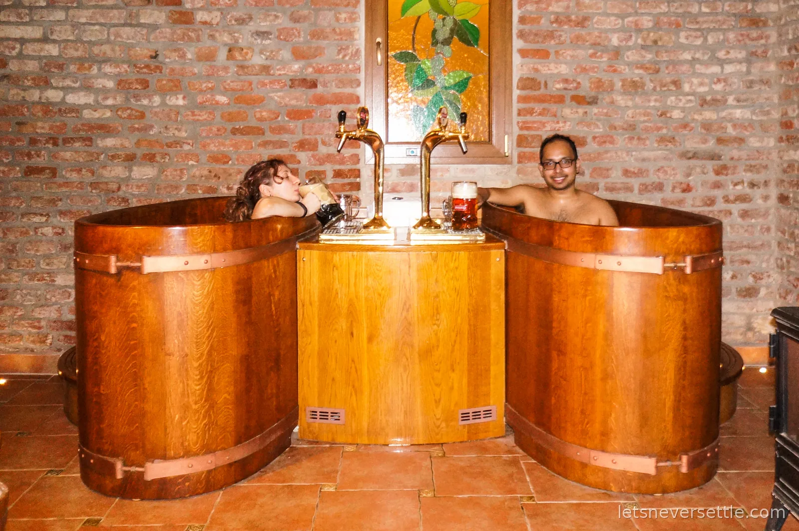 Inga and Dileep in two oak hot tubs in a brick-walled beer spa, each tub with its own pair of beer taps and full glasses