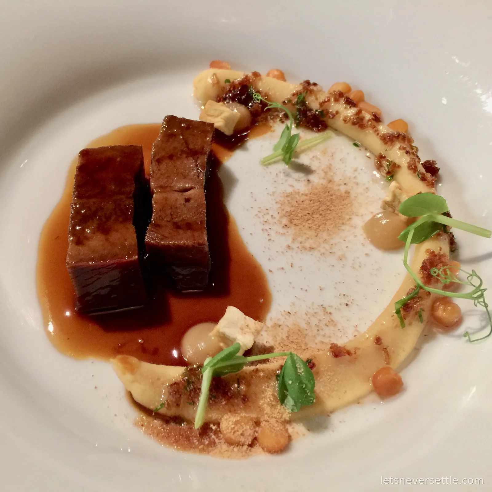 Two glazed cubes of braised beef in dark sauce beside a sweep of puree, crumbs and pea shoots on a white plate