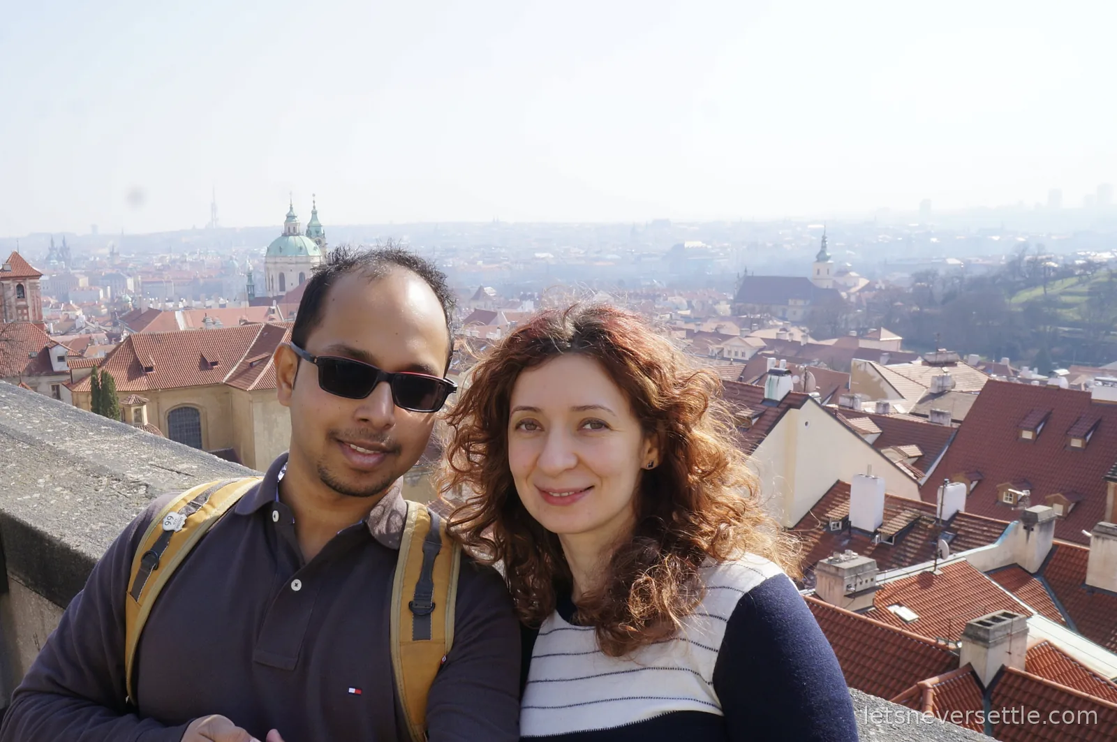 Inga and Dileep on the castle terrace above the red tiled rooftops of Prague, church domes and towers fading into haze behind