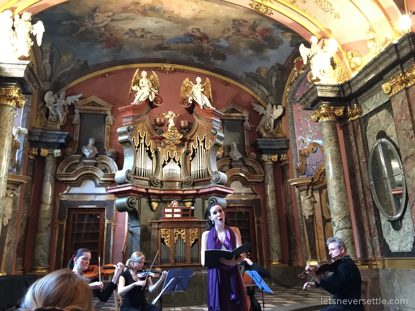 A soprano in a purple dress singing with string players under the frescoed vault and organ of the Mirror Chapel in the Clementinum, Prague