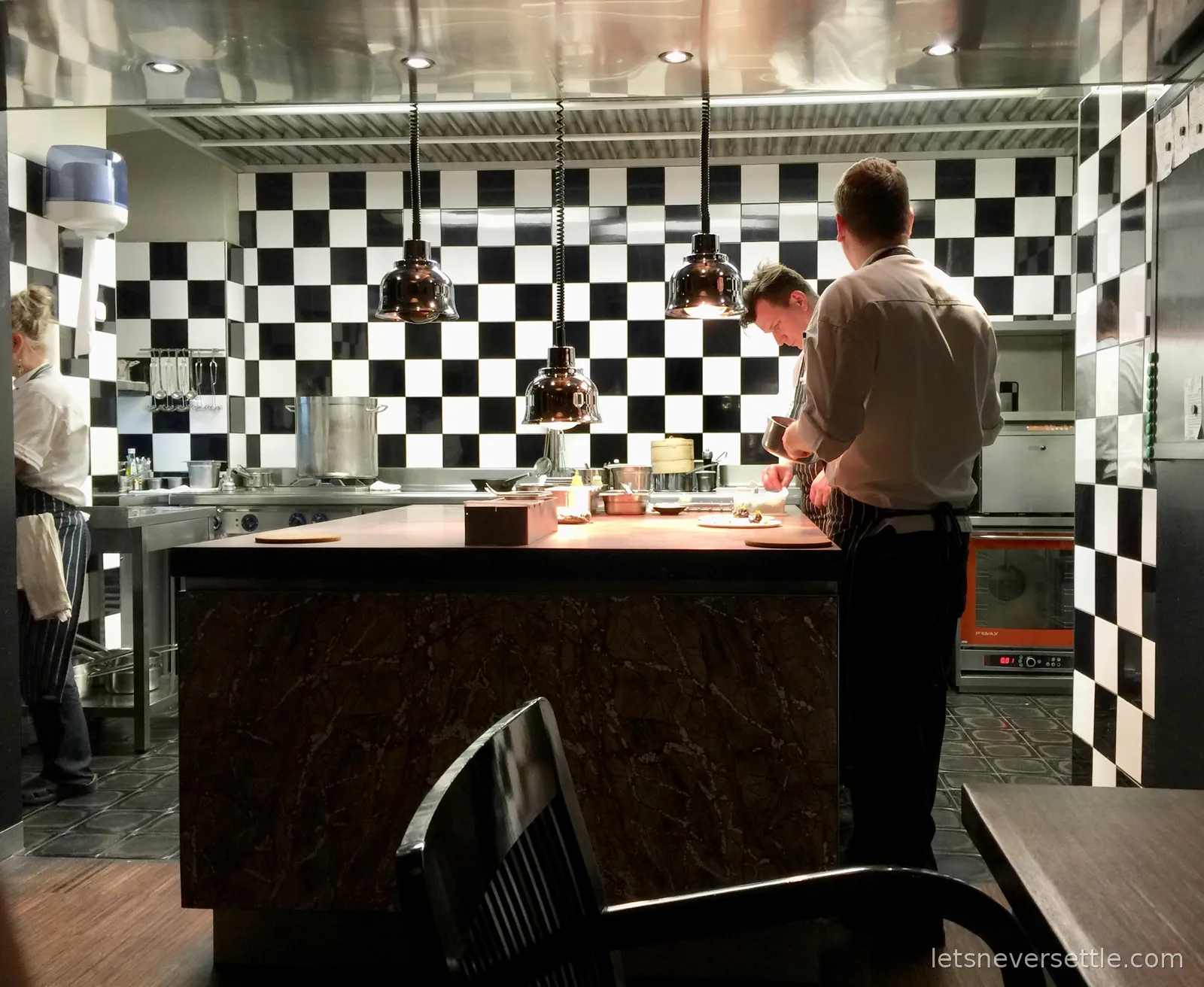The open kitchen at La Degustation in Prague, chefs plating at a marble counter under copper lamps with black and white checkerboard tiles behind them